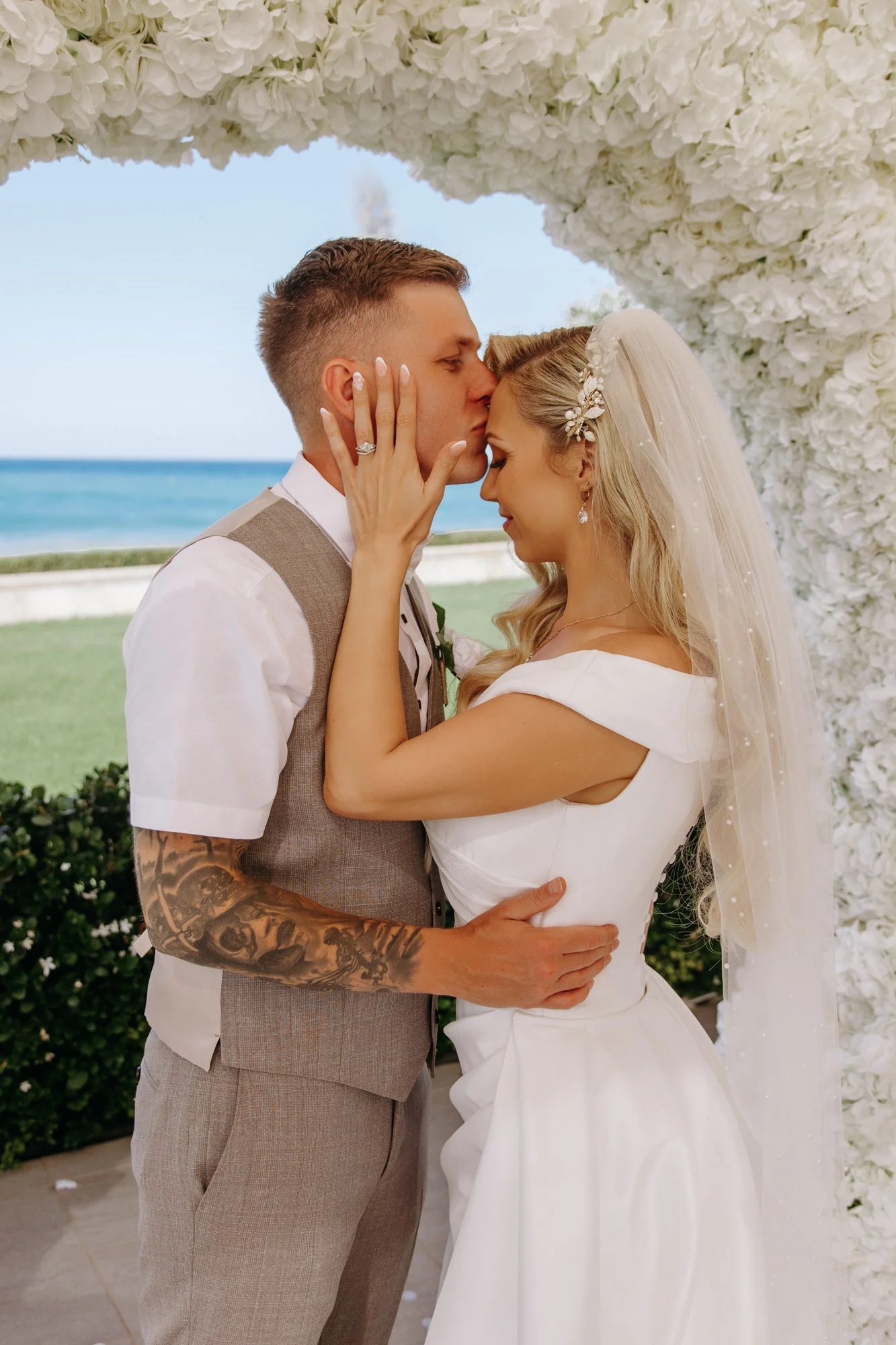 A couple sharing a kiss at their wedding, standing under an arch decorated with white flowers, with the ocean and a grassy area in the background.