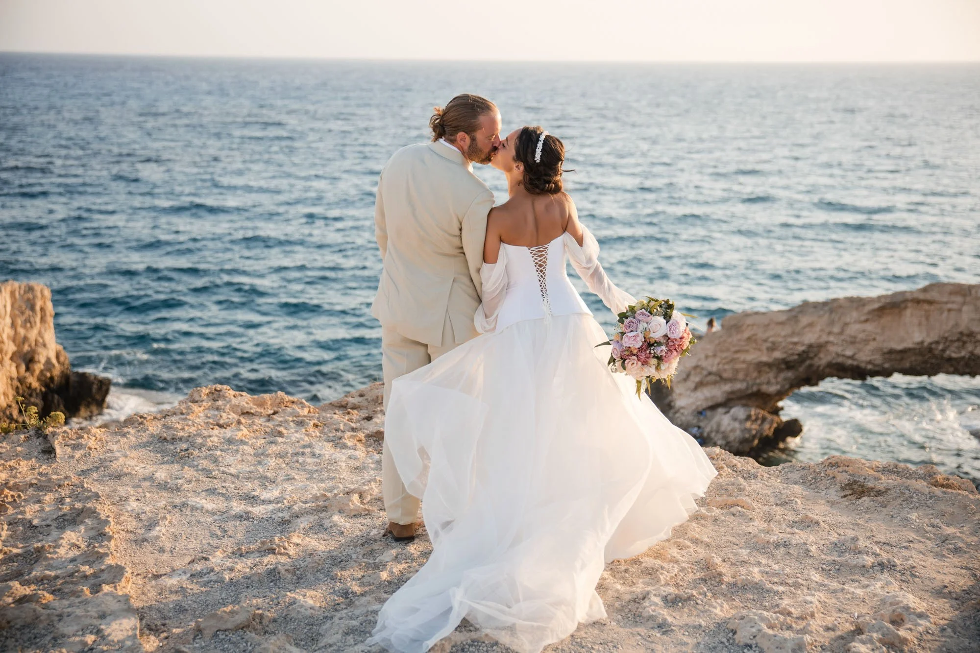 A bride and groom share a kiss by the ocean during their wedding, with rocky shoreline and a large rock arch in the background.