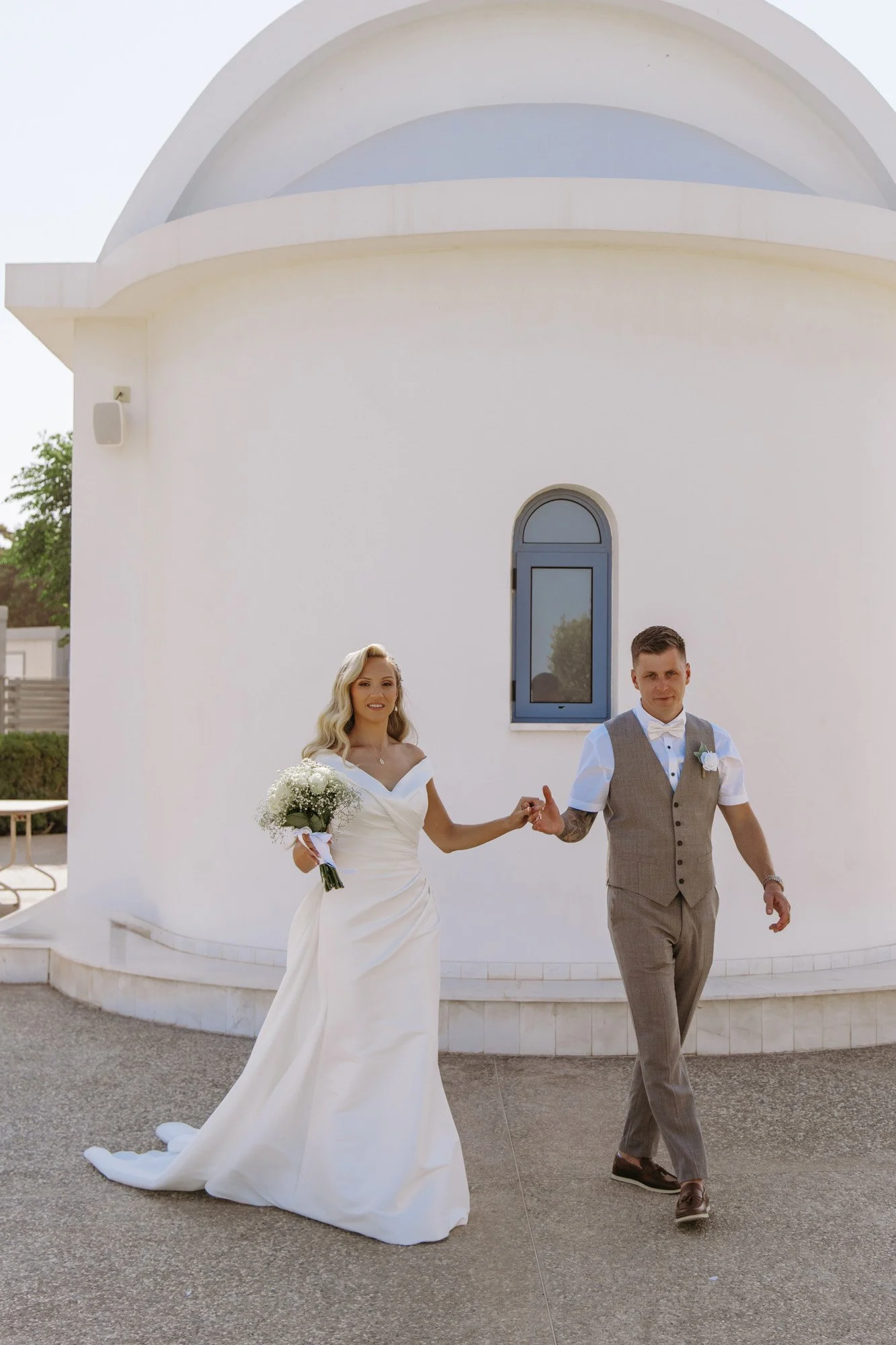 A bride and groom holding hands and walking outside a white chapel or church, with the bride in a white wedding dress holding a bouquet of white flowers and the groom in a gray vest and trousers.