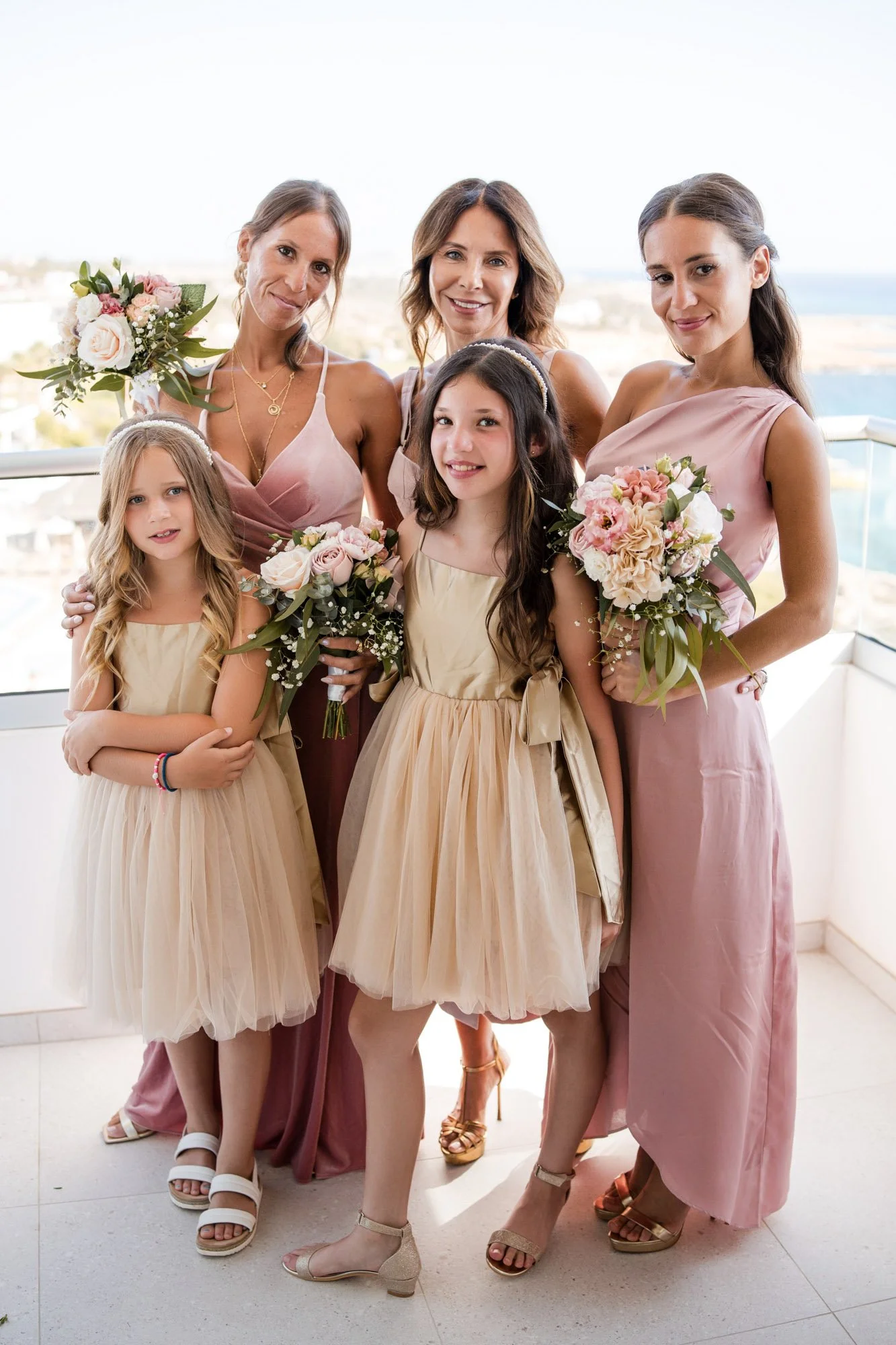 A group of six women and girls dressed in pastel-colored dresses, holding bouquets of flowers, standing on a balcony with a scenic view in the background.