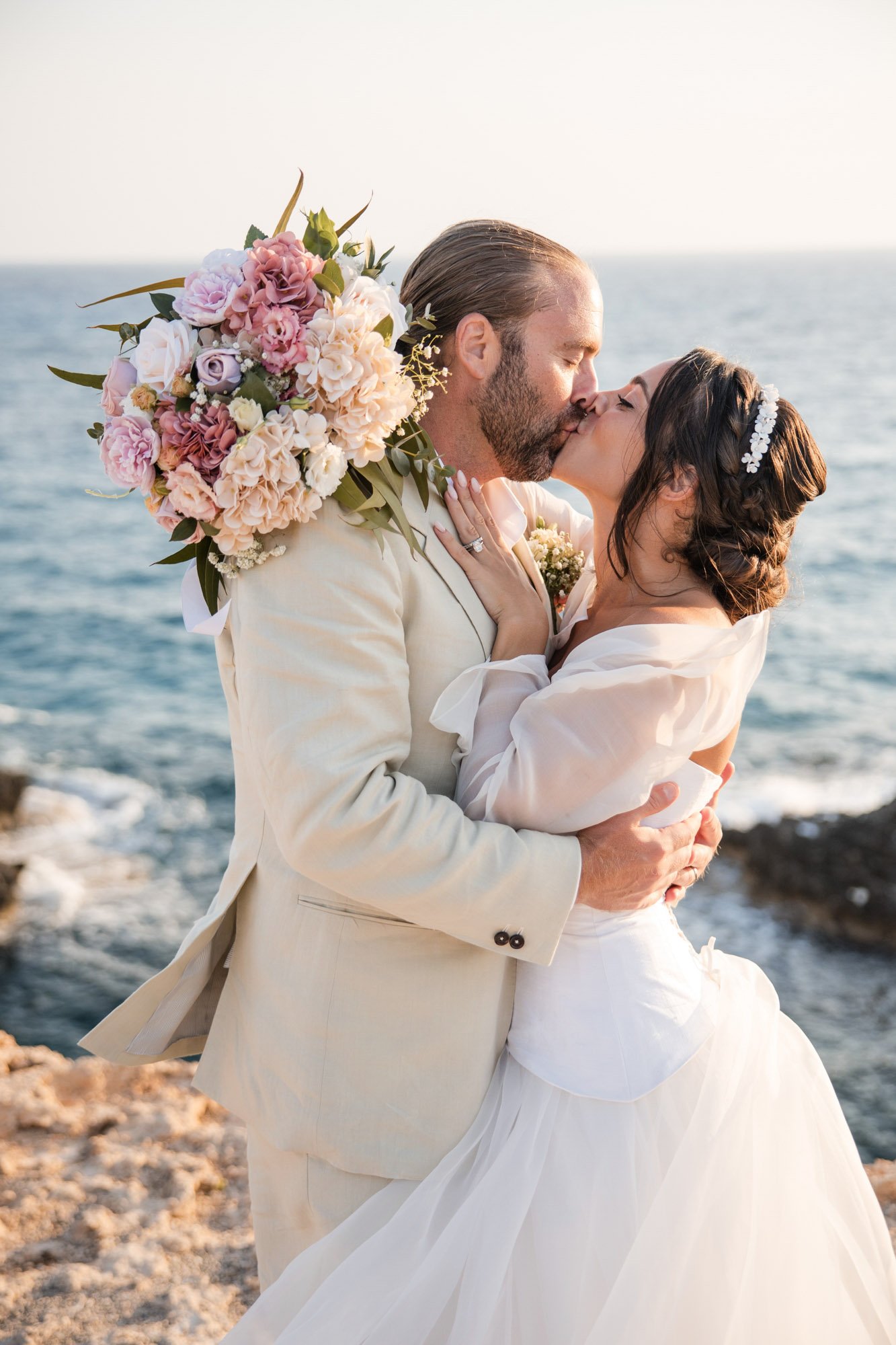 A couple dressed in wedding attire sharing a kiss on a beach, with the ocean in the background and a bouquet of pink and white flowers.