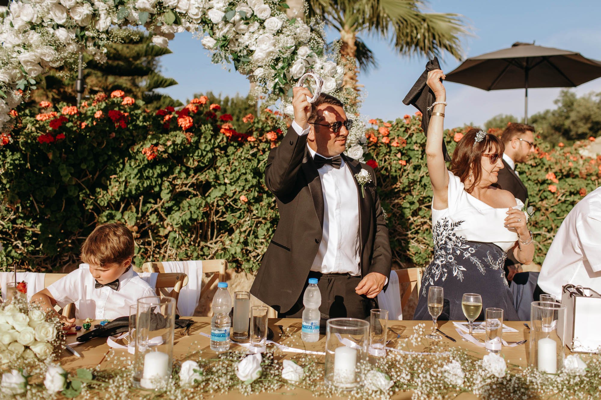 A wedding reception scene with a man in a tuxedo and sunglasses raising his glass, and a woman in an elegant dress and sunglasses raising her hand, both smiling. A young boy in a white shirt and black bow tie is sitting at the table adorned with flow