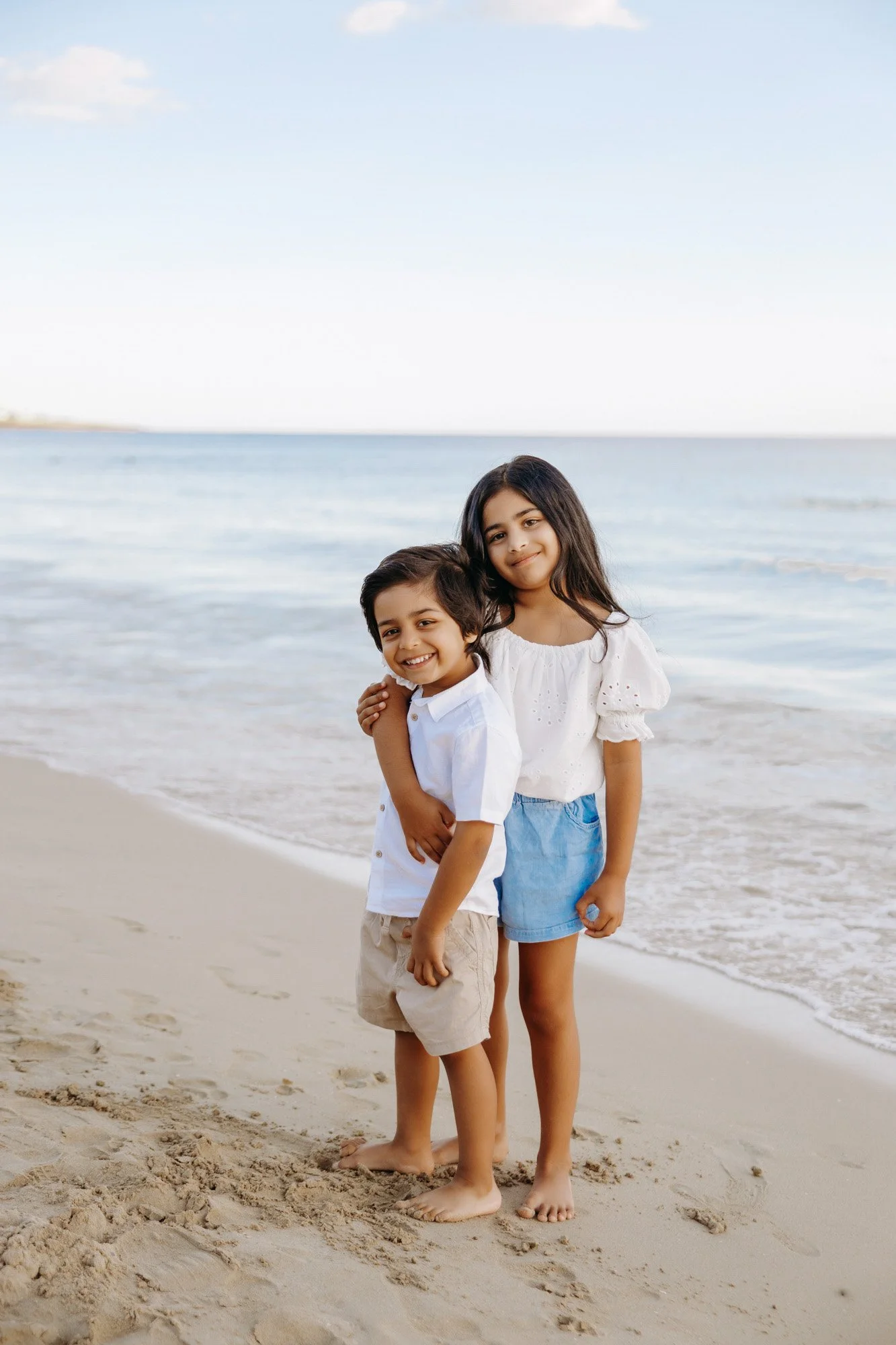 Two children, a boy and a girl, standing on the beach near the water, smiling. The boy is in front, wearing a white shirt and beige shorts, and the girl is behind him, in a white blouse and denim shorts. They are barefoot, with the ocean and sky in t