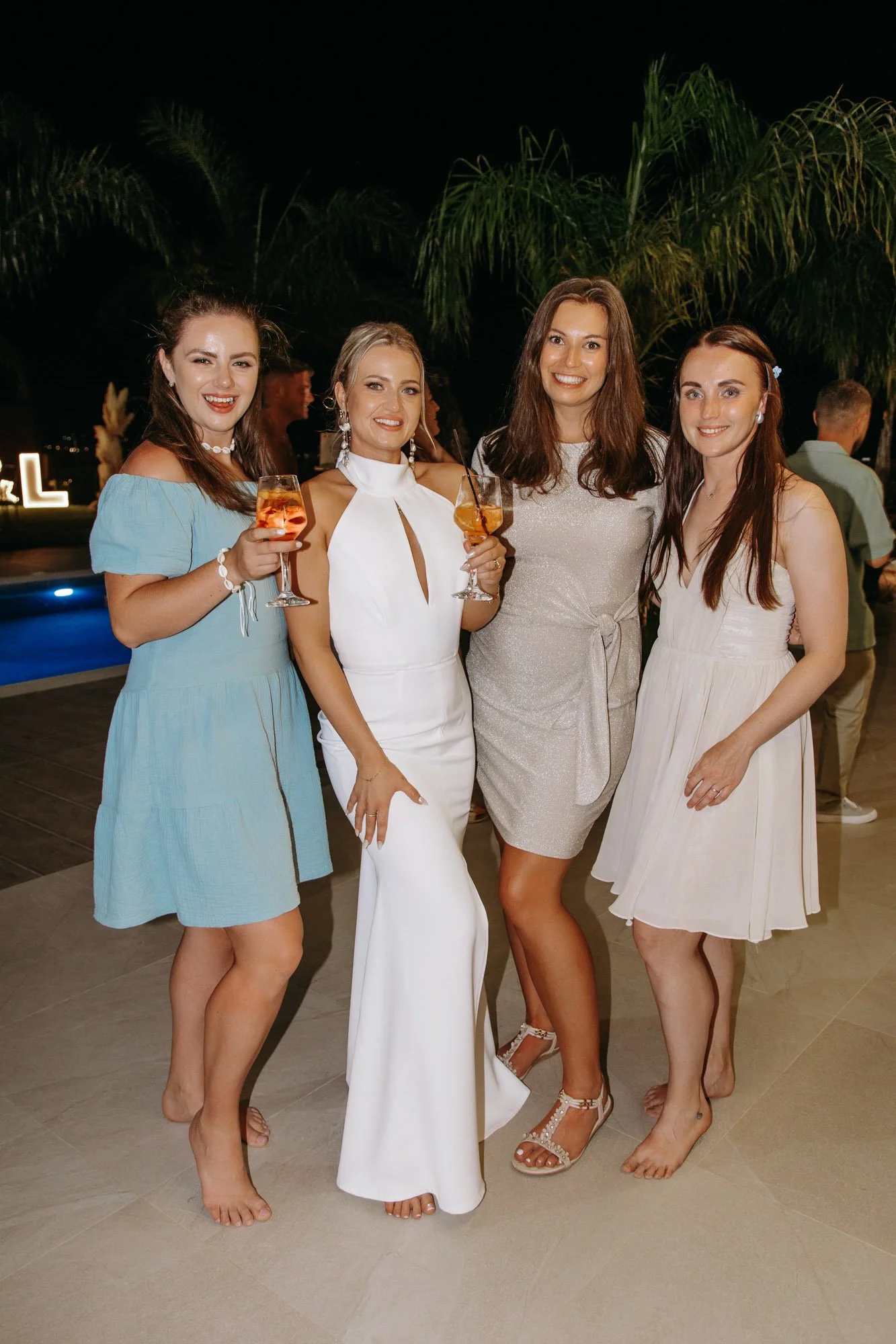 Group of four women in party dresses, smiling, holding drinks, at an outdoor evening event near a pool with palm trees in the background.