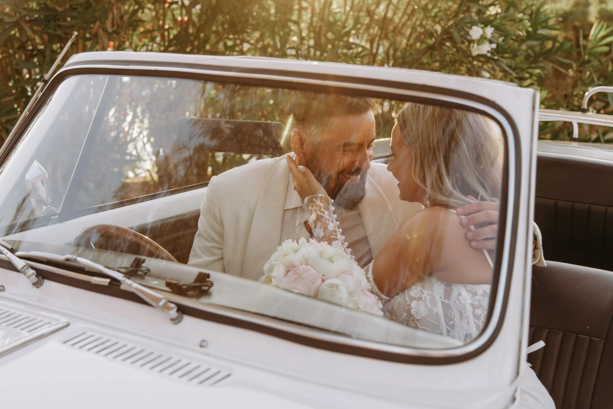A man and woman dressed in wedding attire, sitting in a vintage white convertible car, sharing a joyful moment together, with the woman holding a bouquet of flowers.