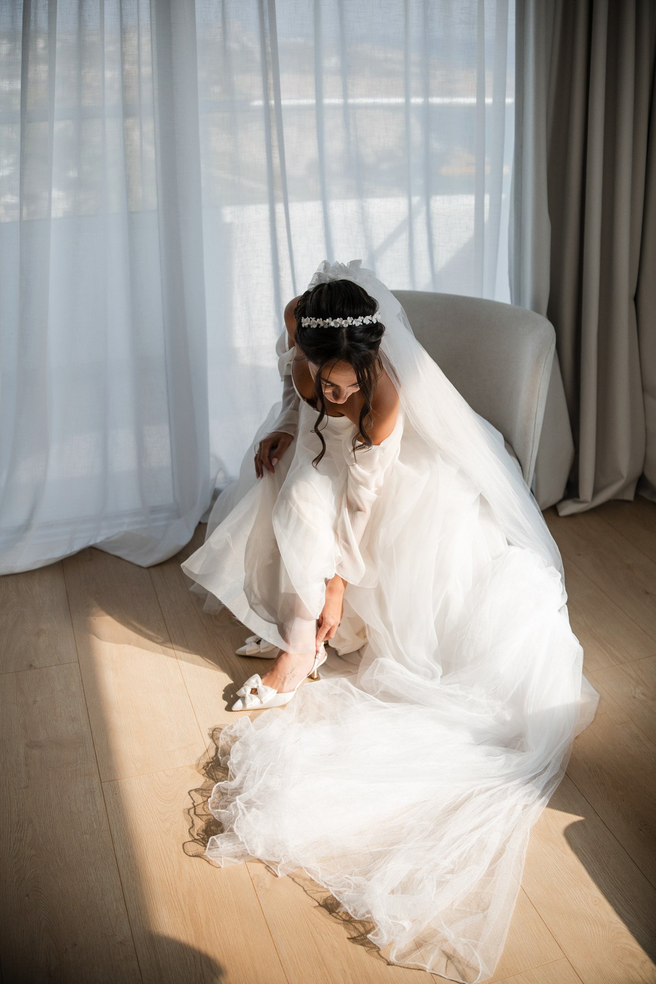 A bride in a white wedding gown sitting on a chair, wearing a tiara, adjusting her high-heeled shoe in a room with sheer curtains and sunlight coming through.