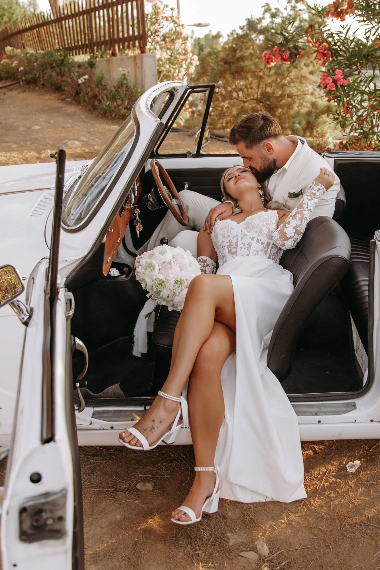 A bride and groom in wedding attire sitting in a vintage convertible car; the groom is kissing the bride on the forehead, and she is leaning back with her eyes closed, holding a bouquet of flowers.