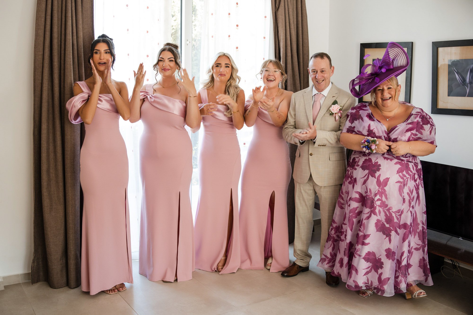 Group of six women and one man dressed in pink and beige attending a wedding, posing indoors near a window with curtains, smiling and celebrating.