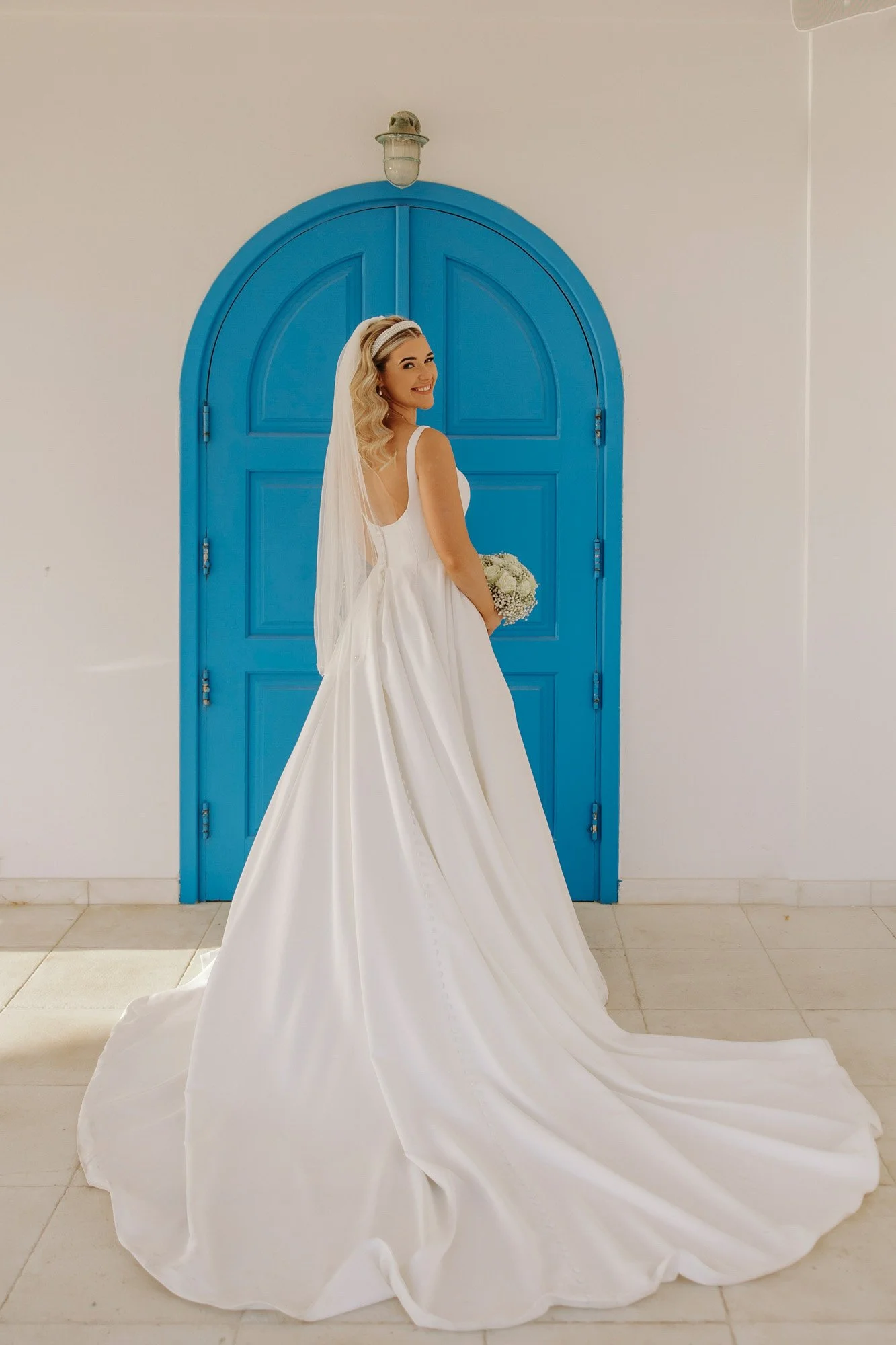 Bride in a white wedding dress holding a bouquet, standing in front of a bright blue arched door with a wall-mounted light fixture above.
