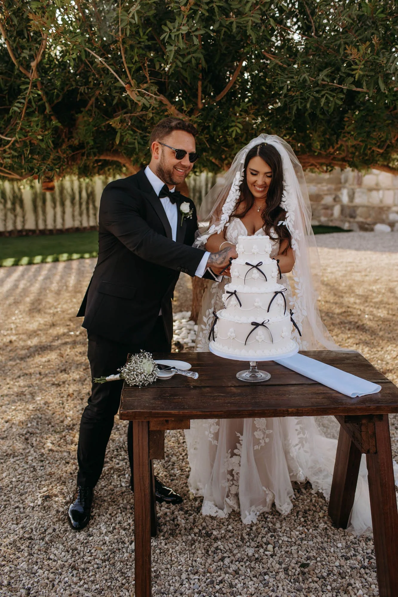 A newlywed couple cutting a wedding cake outdoors under a tree. The groom is wearing a black tuxedo and sunglasses, and the bride is in a lace wedding dress with a veil. The cake is white with black ribbon decorations.