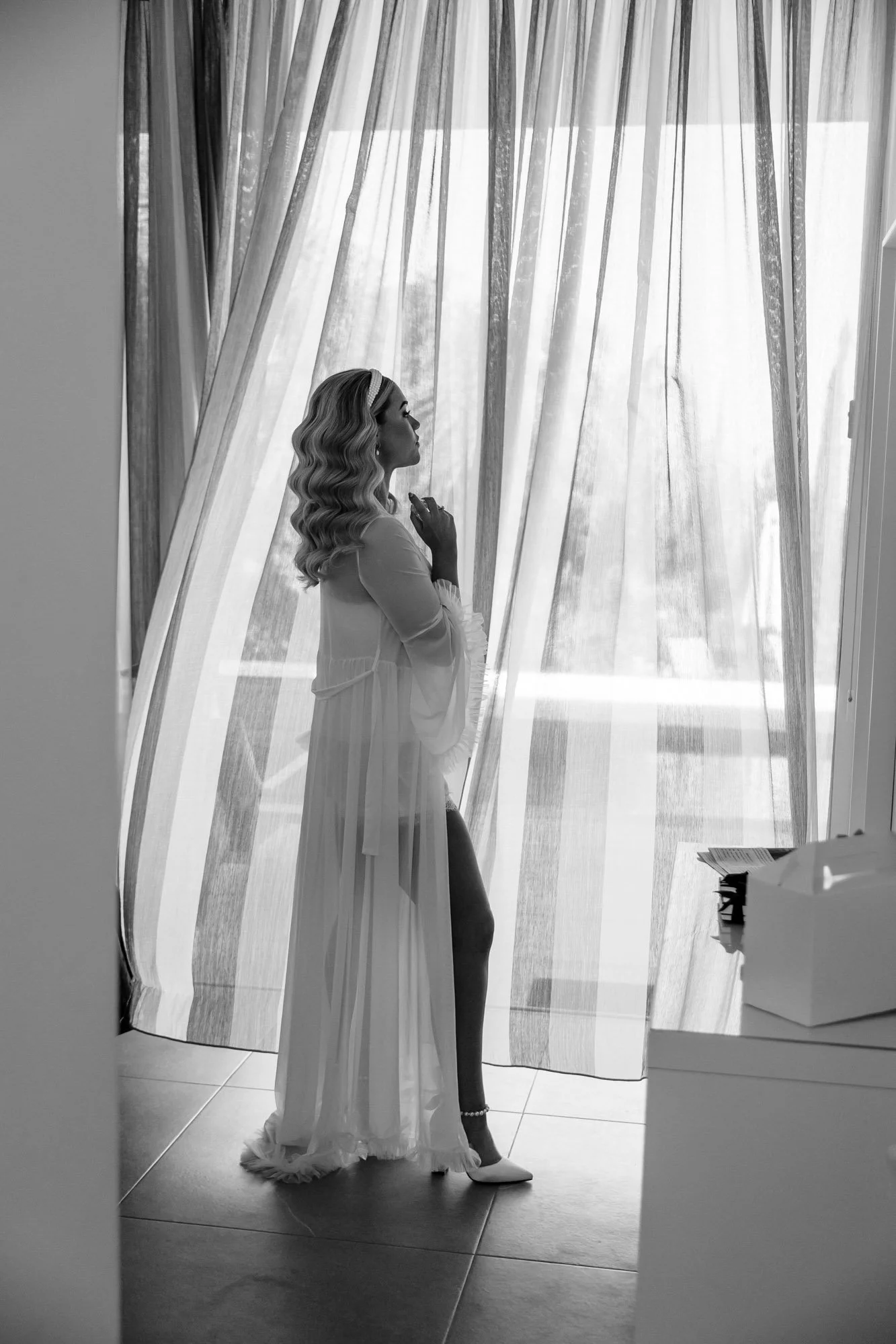 A woman with styled blonde hair, dressed in a sheer white robe and high heels, stands near a window with curtains, looking pensively outside.