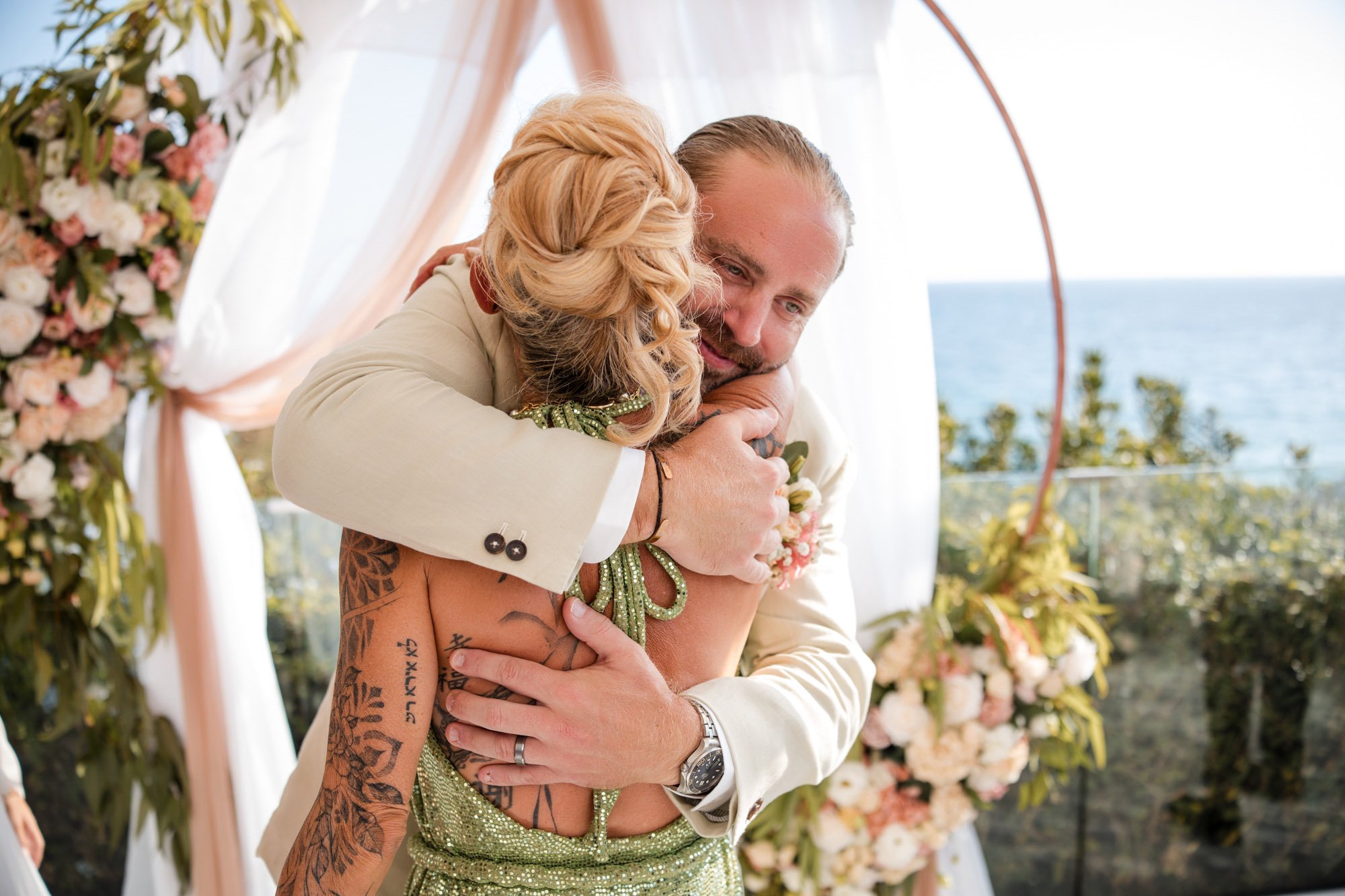 A couple hugging at their wedding ceremony outdoors with an ocean view in the background. The bride has tattoos and is wearing a green dress, and the groom is in a white suit. They are embracing warmly near floral decorations.
