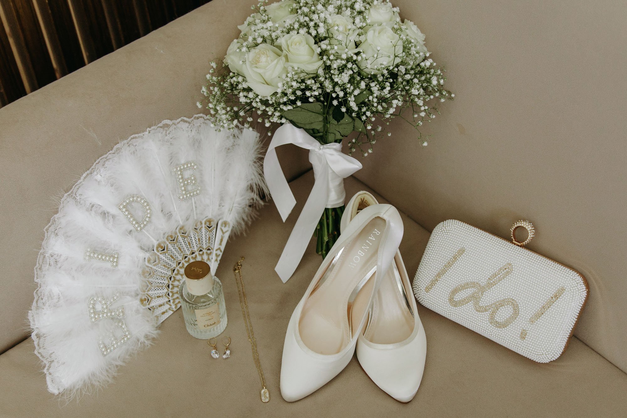 Wedding accessories including a bouquet of white roses and baby's breath, white high heels, a jewelry necklace and earrings, a pearl-embellished clutch with the word "I do!" and a bottle of perfume, all arranged on a beige sofa.