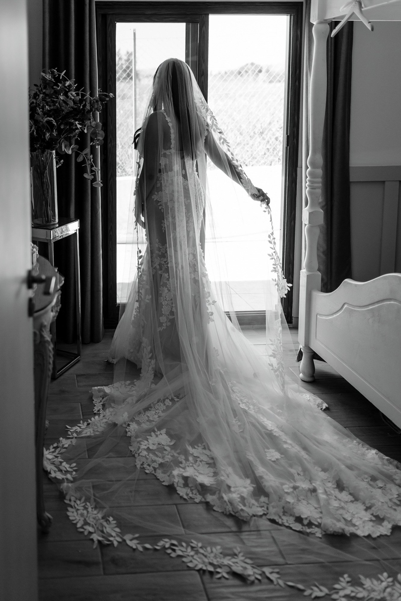 A woman in a wedding dress with a long train and veil, standing near a sliding glass door with sunlight filtering in.