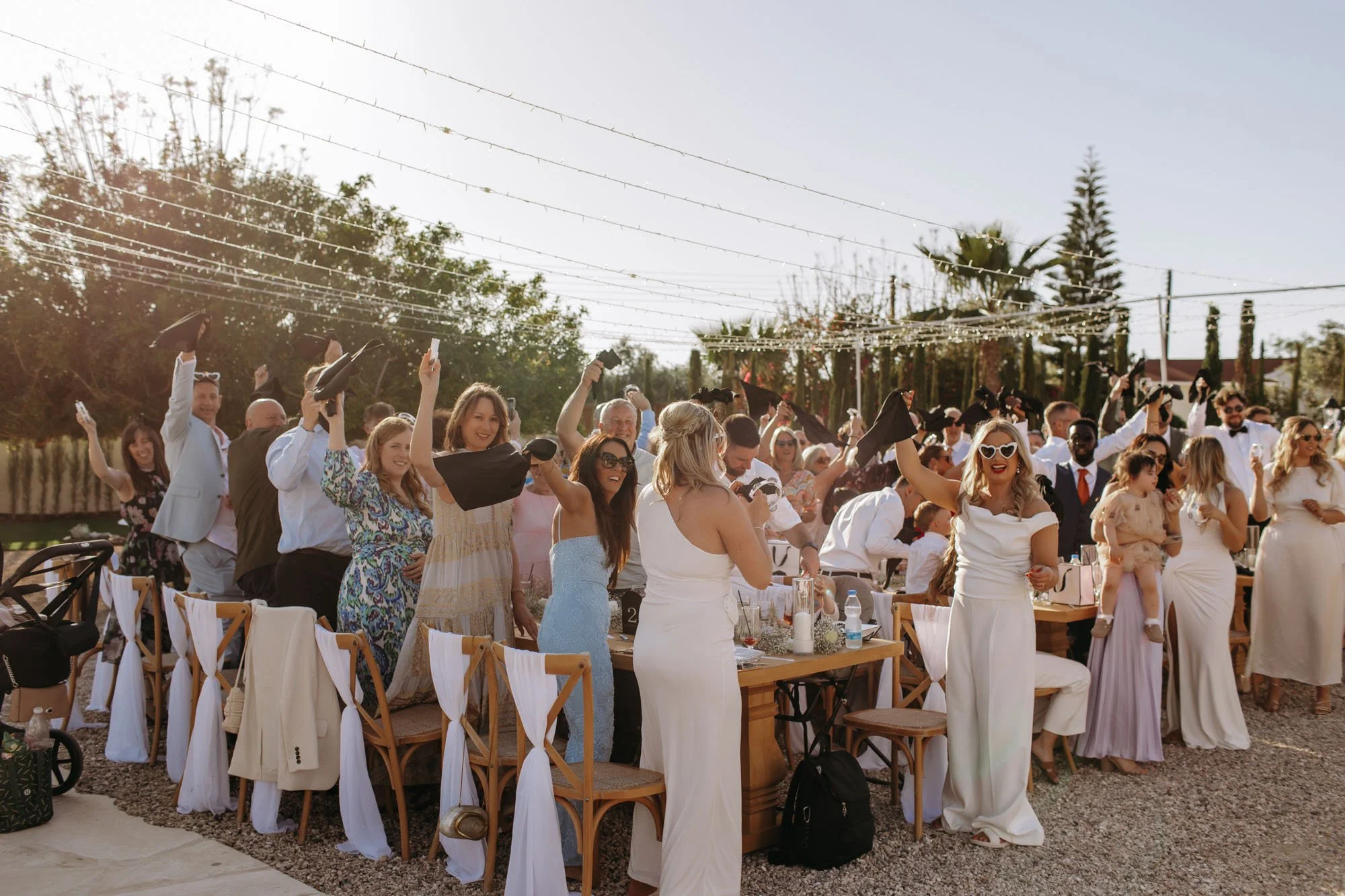 People celebrating at an outdoor wedding reception, some wearing graduation caps, with tables, chairs, and string lights overhead.