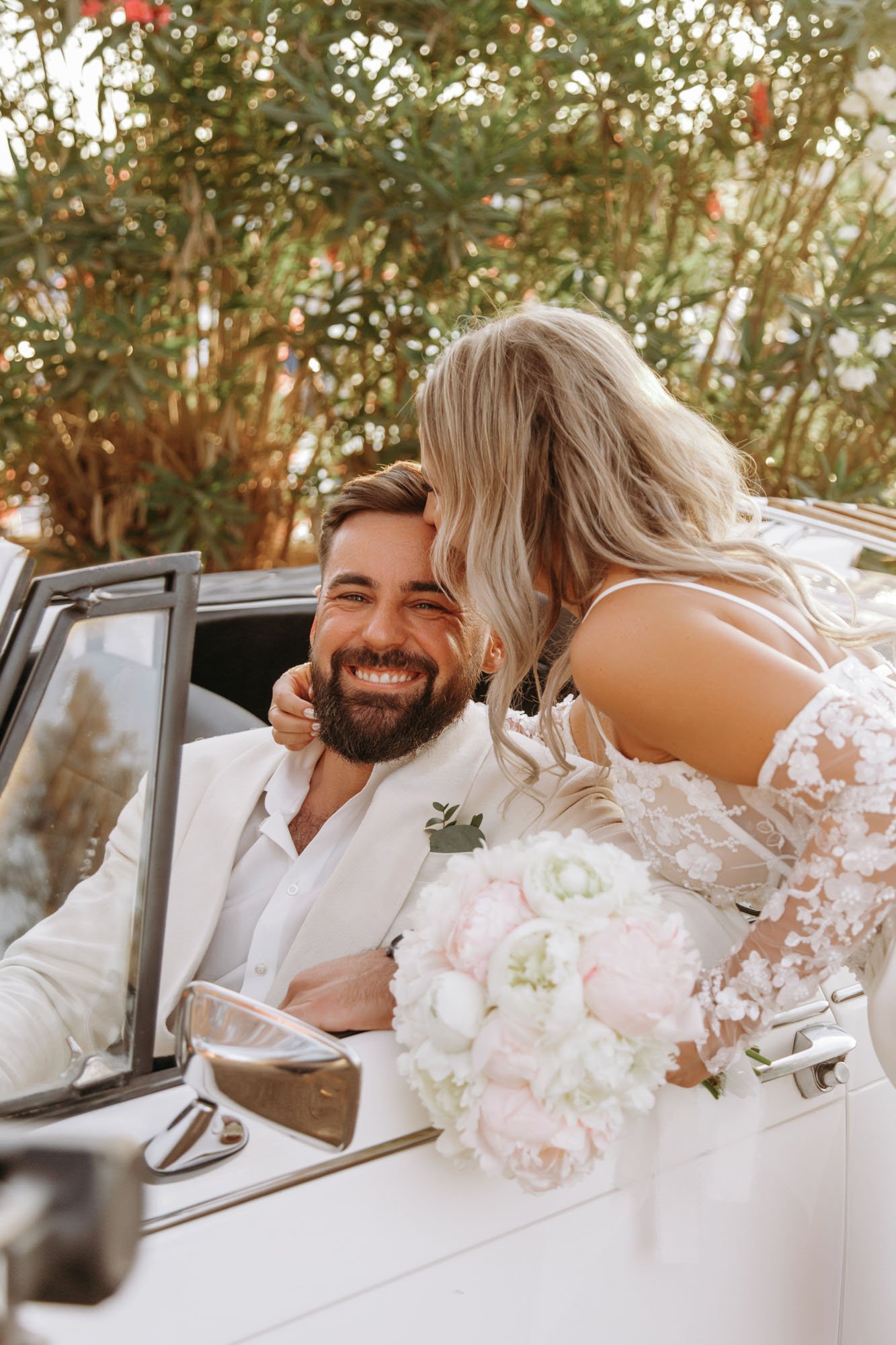 A newlywed couple in wedding attire sitting in a vintage convertible car, with the bride giving the groom a kiss on the forehead. The groom is smiling and holding a bouquet of pink and white flowers. They are outdoors with greenery in the background.