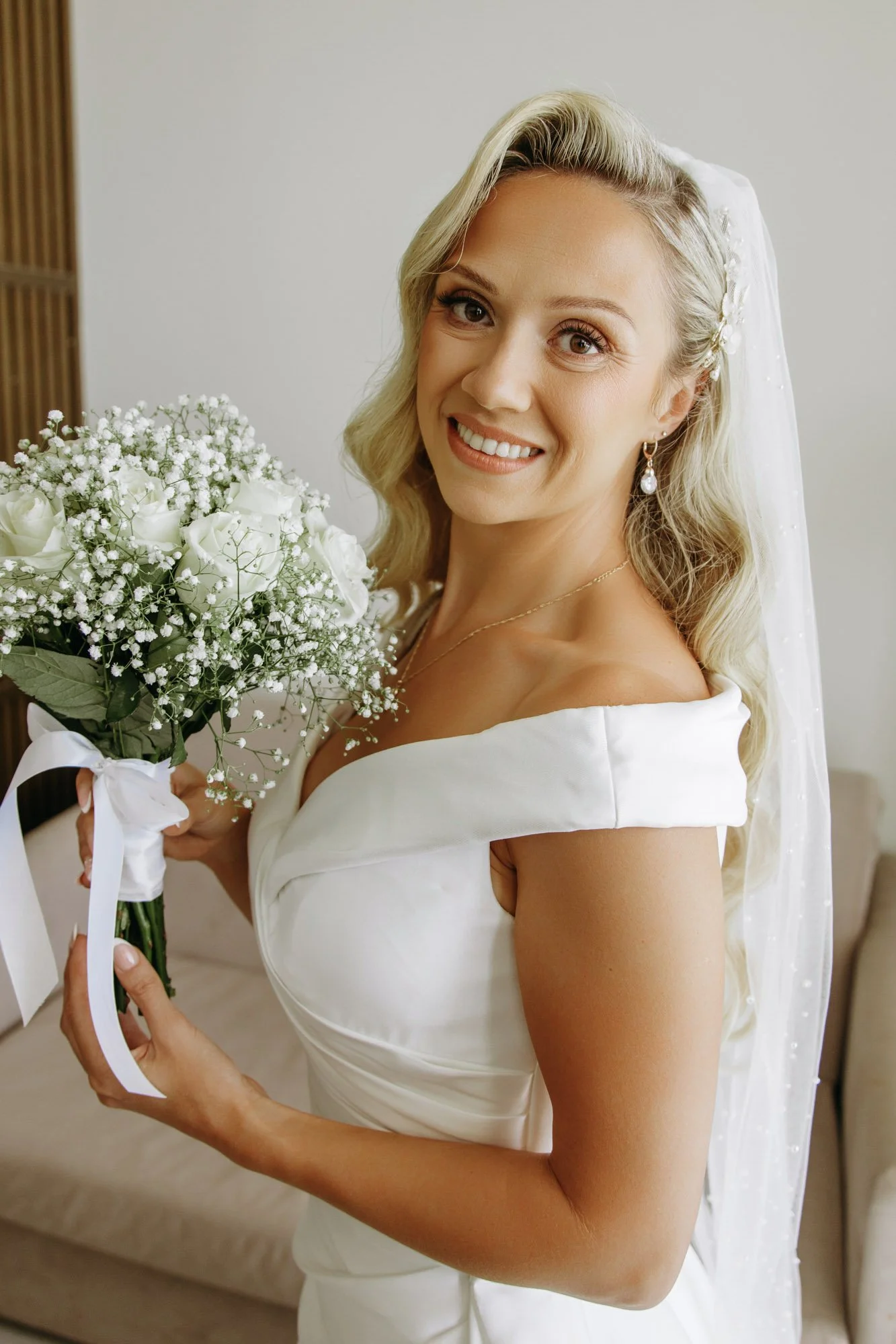 A smiling bride with blonde hair holding a bouquet of white roses and baby's breath, wearing a white wedding gown with off-shoulder sleeves and a veil.