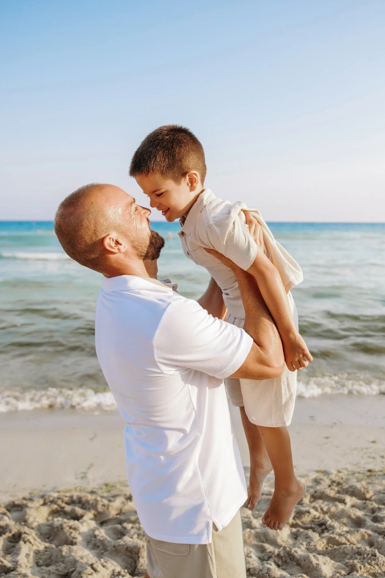 A man is holding a boy in his arms at the beach, both smiling and looking into each other's eyes, with the ocean and a clear sky in the background.