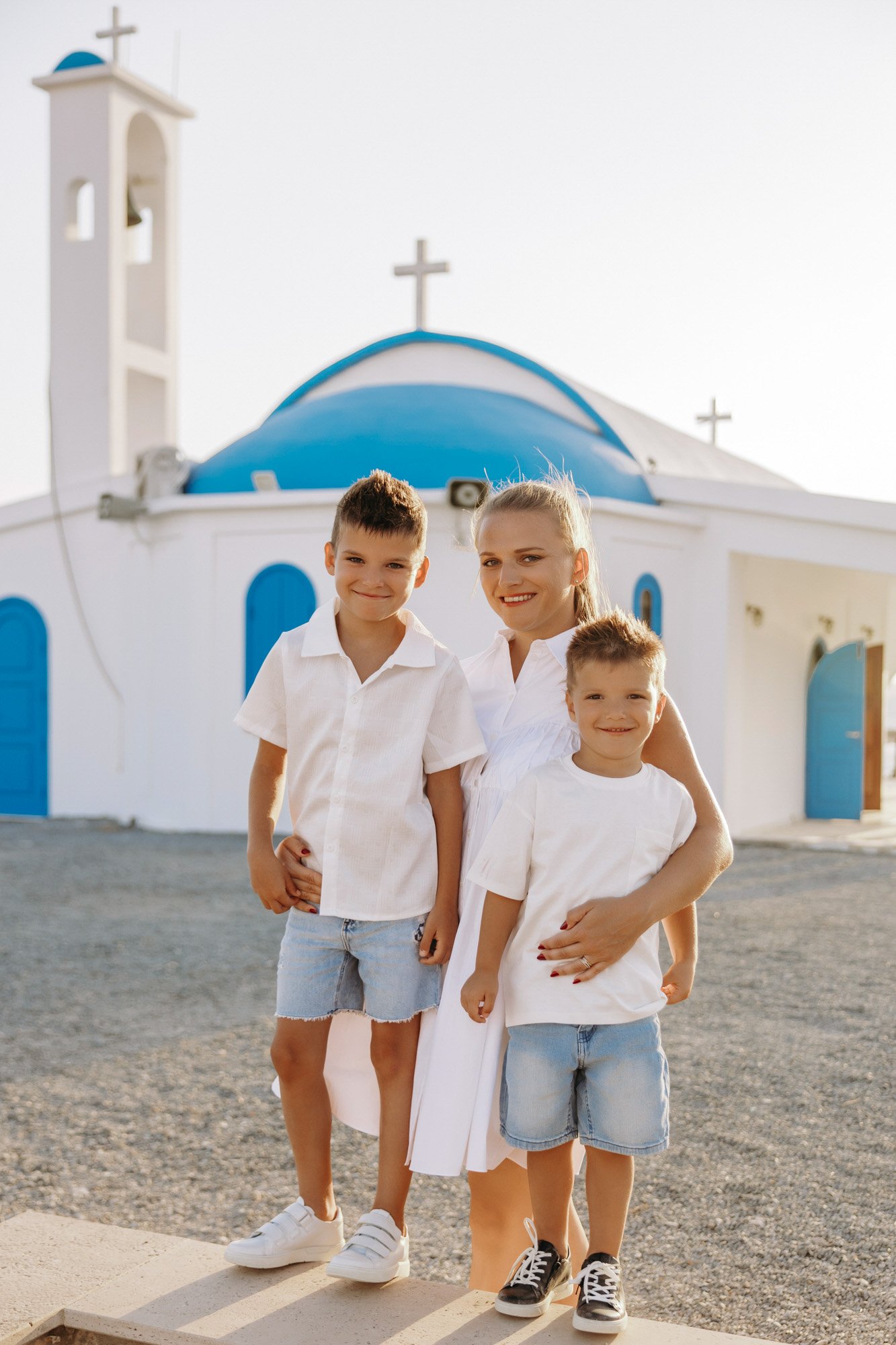 A woman and three children stand in front of a white and blue church with crosses on the roof and bell tower, during daytime.