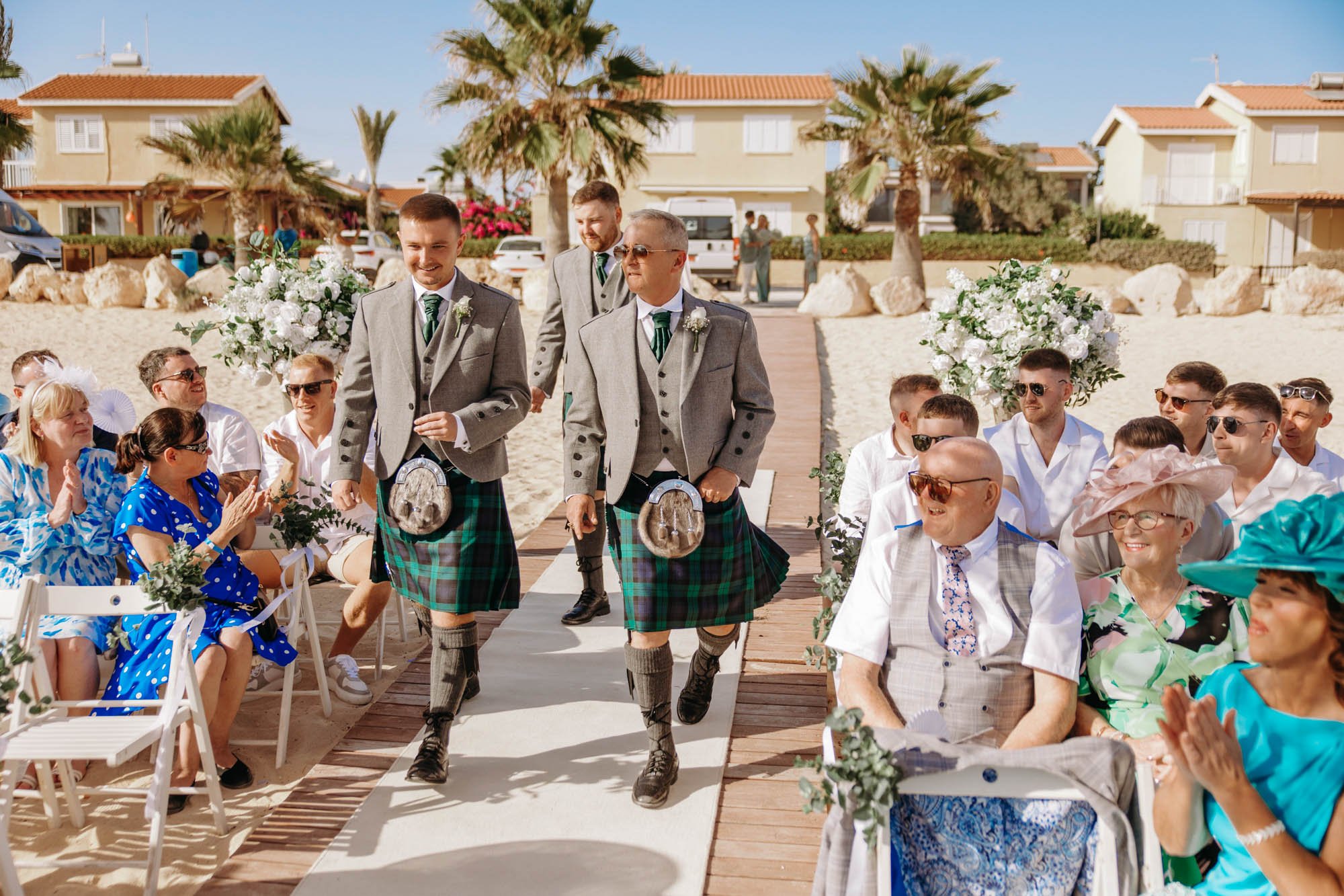 Groomsmen dressed in gray suits and kilts walking down an outdoor aisle at a beach wedding, with seated guests clapping and smiling.