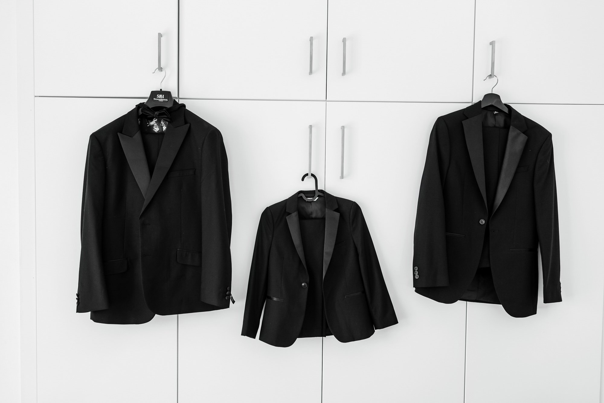 Three black tuxedo jackets hung on hooks against a white cabinet wall.
