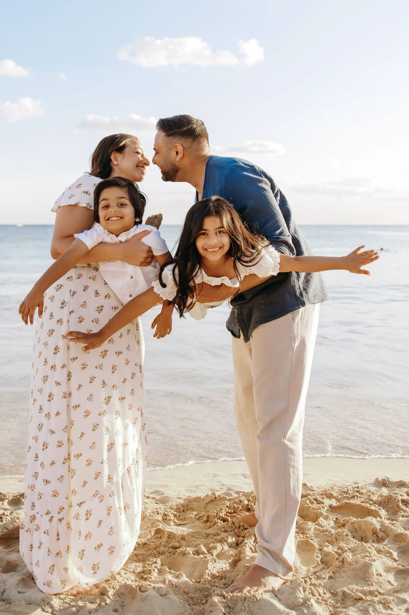 Family of four on the beach, with a woman and man leaning toward each other, smiling, while two children pose playfully, surrounded by sand and ocean waves.