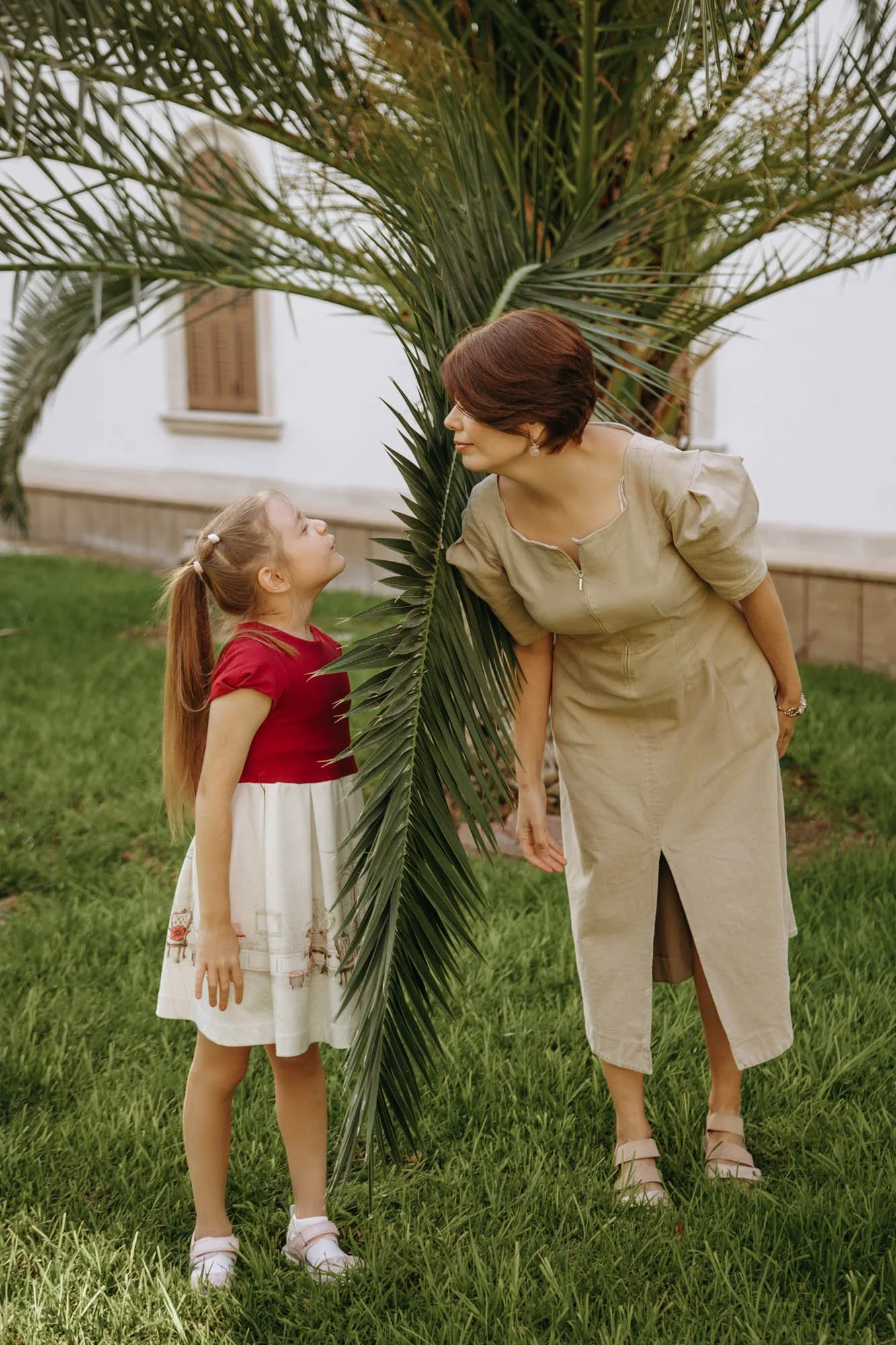 A woman and a young girl look at each other behind a large palm tree on a grassy lawn outside a house with white walls and brown window shutters.