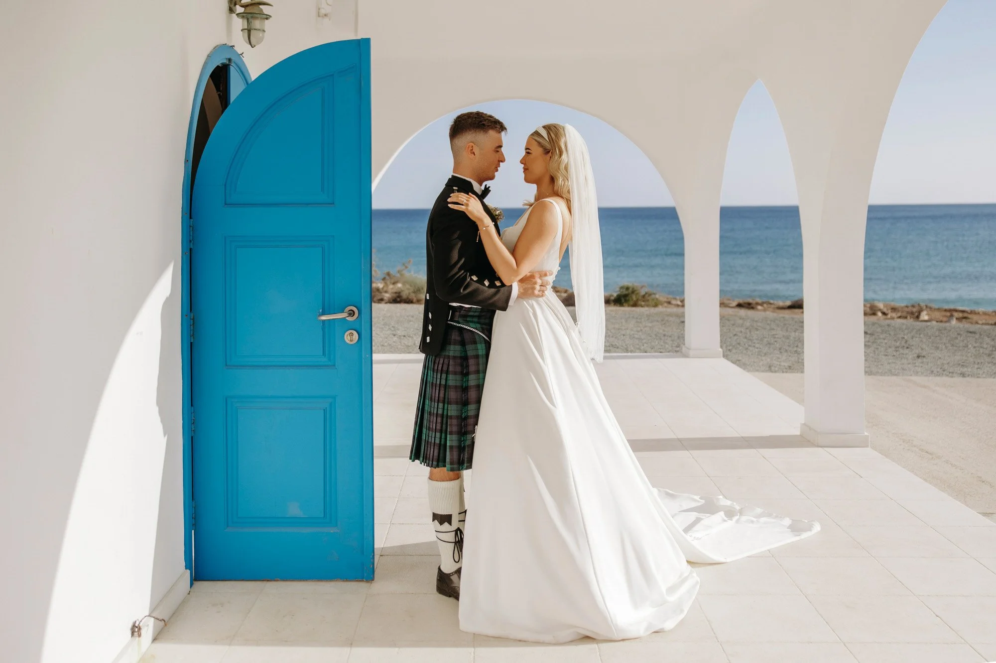A bride and groom standing close together outdoors near the ocean, with a blue door and white arches in the background, during their wedding ceremony.