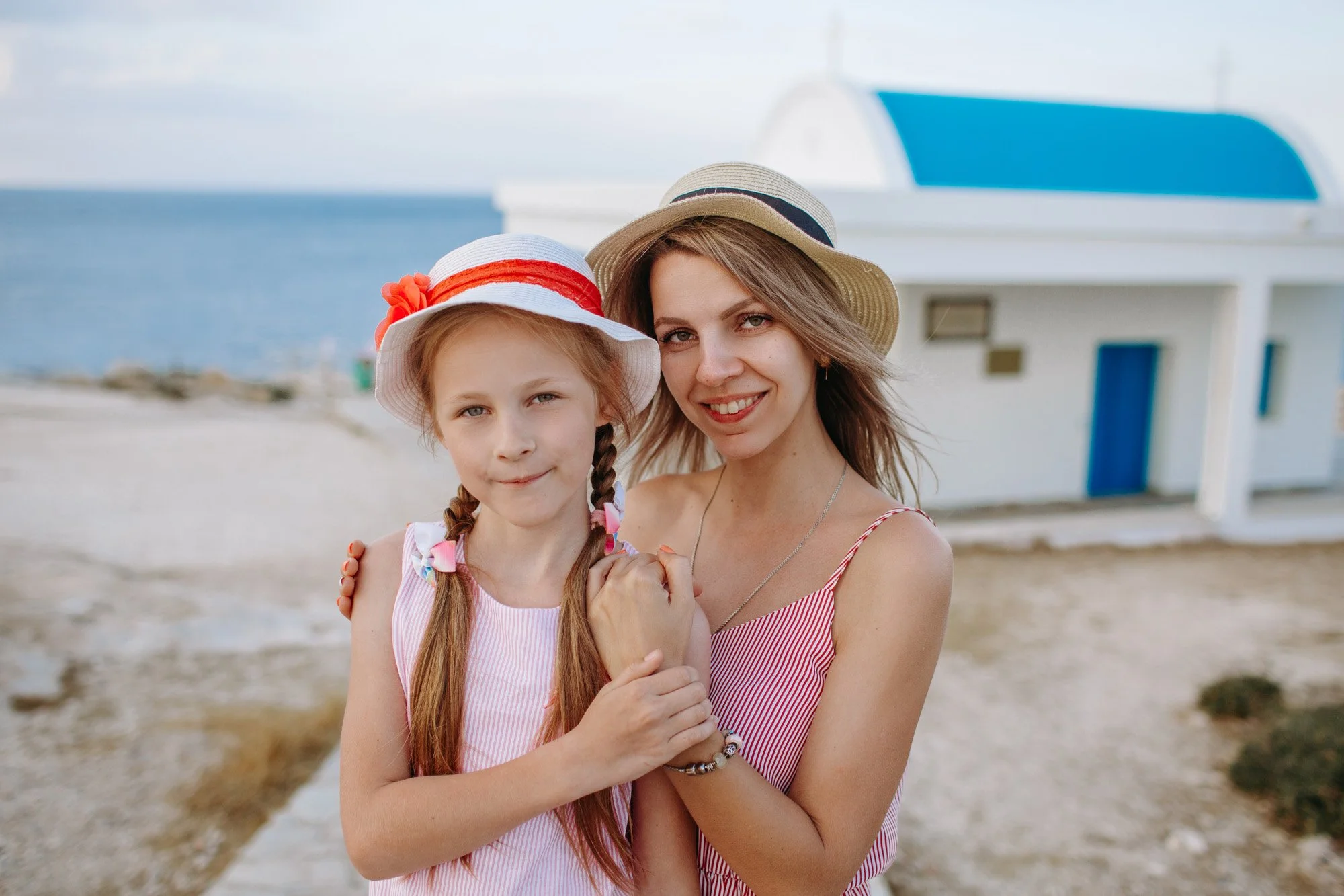 A woman and young girl share a hug on a beach, both wearing summer dresses and straw hats, with the ocean and a white coastal building in the background.