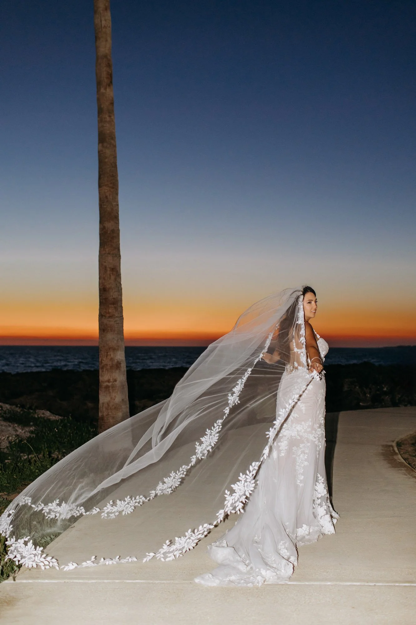 A woman in a white wedding dress and veil standing on a path near the ocean during sunset.