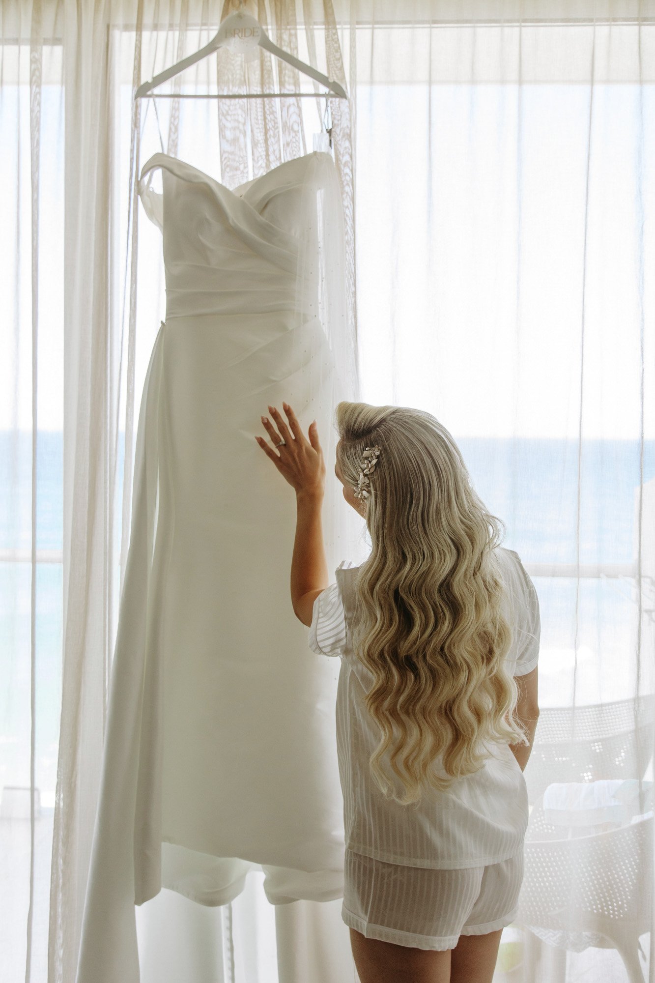 A woman with long, wavy blonde hair and a decorative hair clip is looking at a white wedding dress hanging on a hanger. The scene is backlit by sheer curtains, with a view of the ocean in the background.