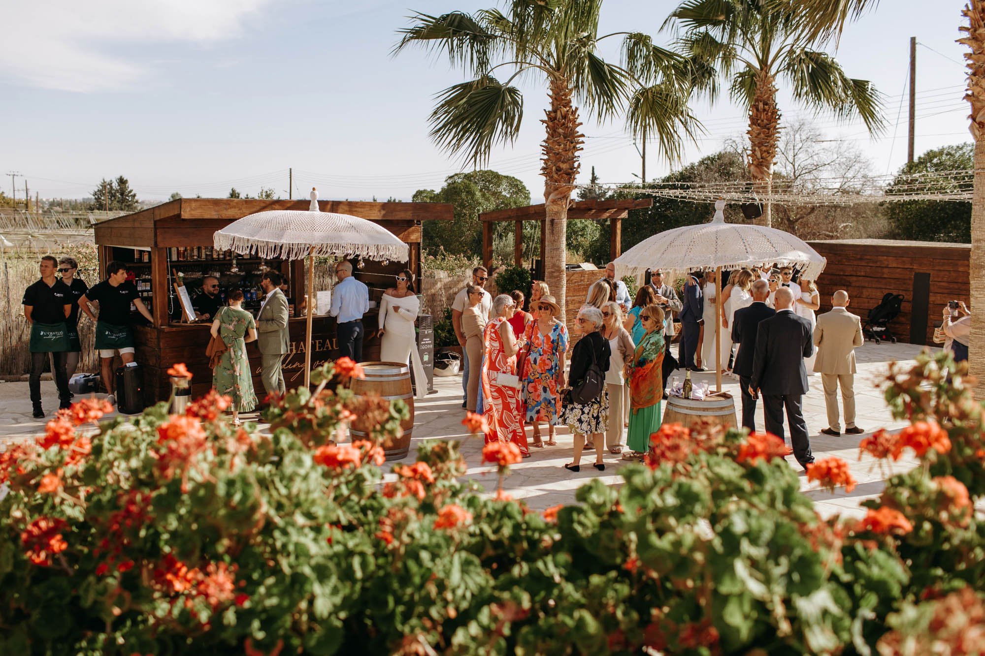 People socializing at an outdoor event under large white parasols, with a wooden bar, palm trees, and flowers in the foreground.