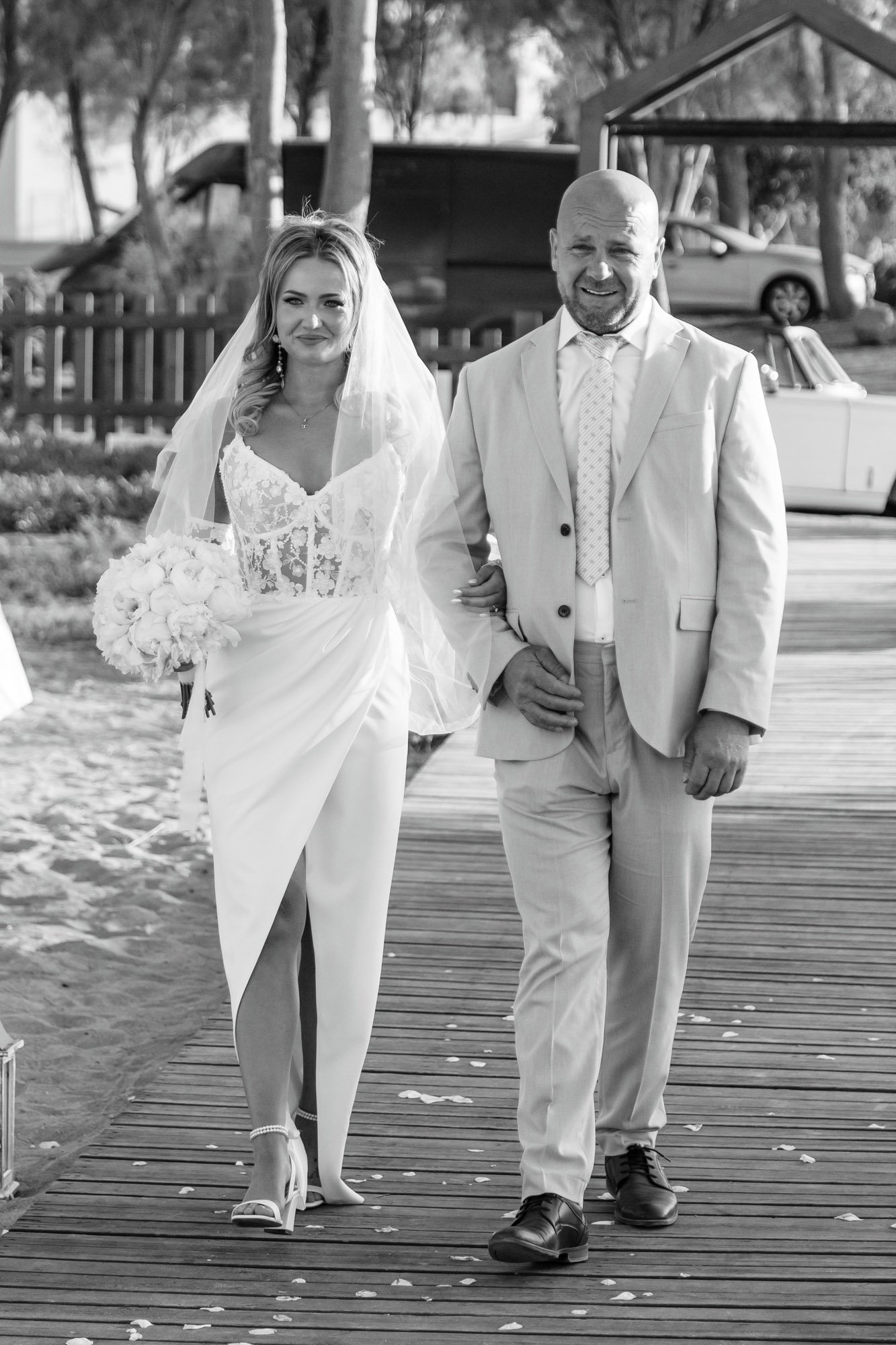 A bride in a white wedding dress and veil walks arm-in-arm with an older man in a light-colored suit on a wooden pathway outdoors, with flowers on the ground around them, trees, and parked cars in the background.