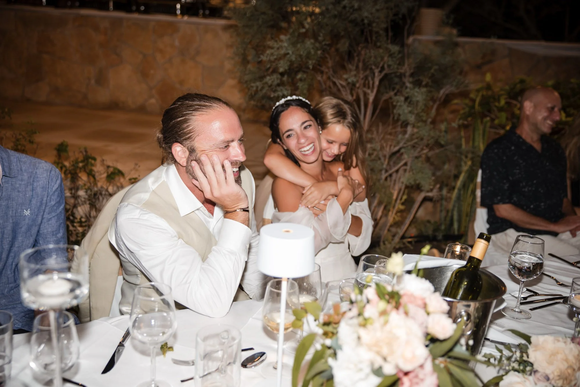 People sitting at a dinner table, laughing and enjoying a celebration, with floral centerpieces, wine bottles, and glasses.