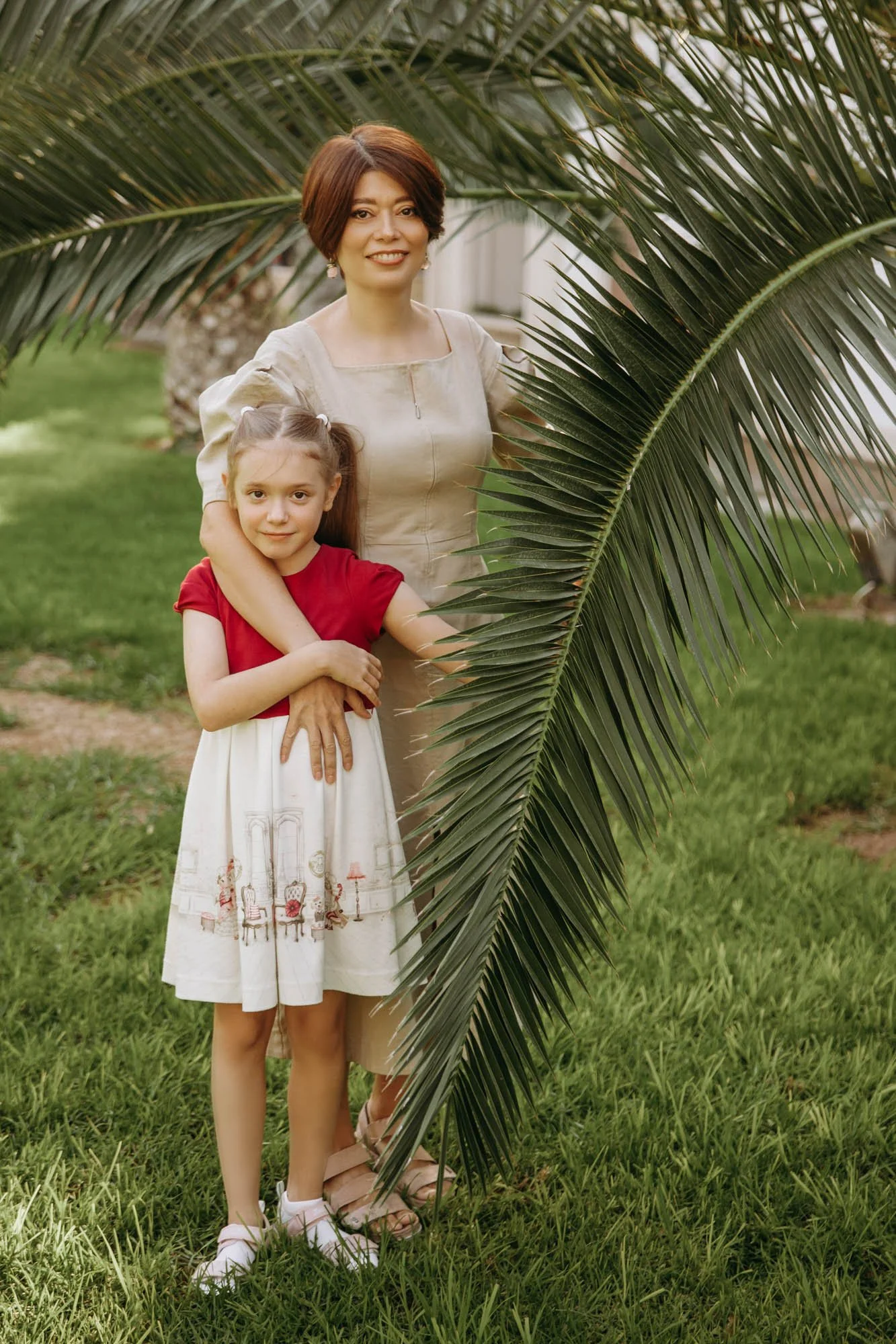A woman with short brown hair and a girl with long brown hair standing outdoors behind a large palm frond. The woman is wearing a beige dress and the girl is wearing a red top and white skirt with a scene of chairs and lamps. They are on green grass 