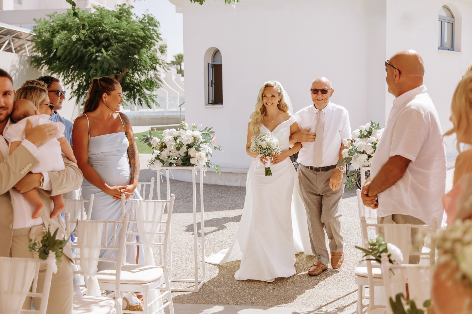 A bride in a white wedding dress walking arm-in-arm with an older man, likely her father, at an outdoor wedding ceremony. Guests, including children and adults, stand on either side, dressed in light-colored and pastel clothing. White floral arrangem