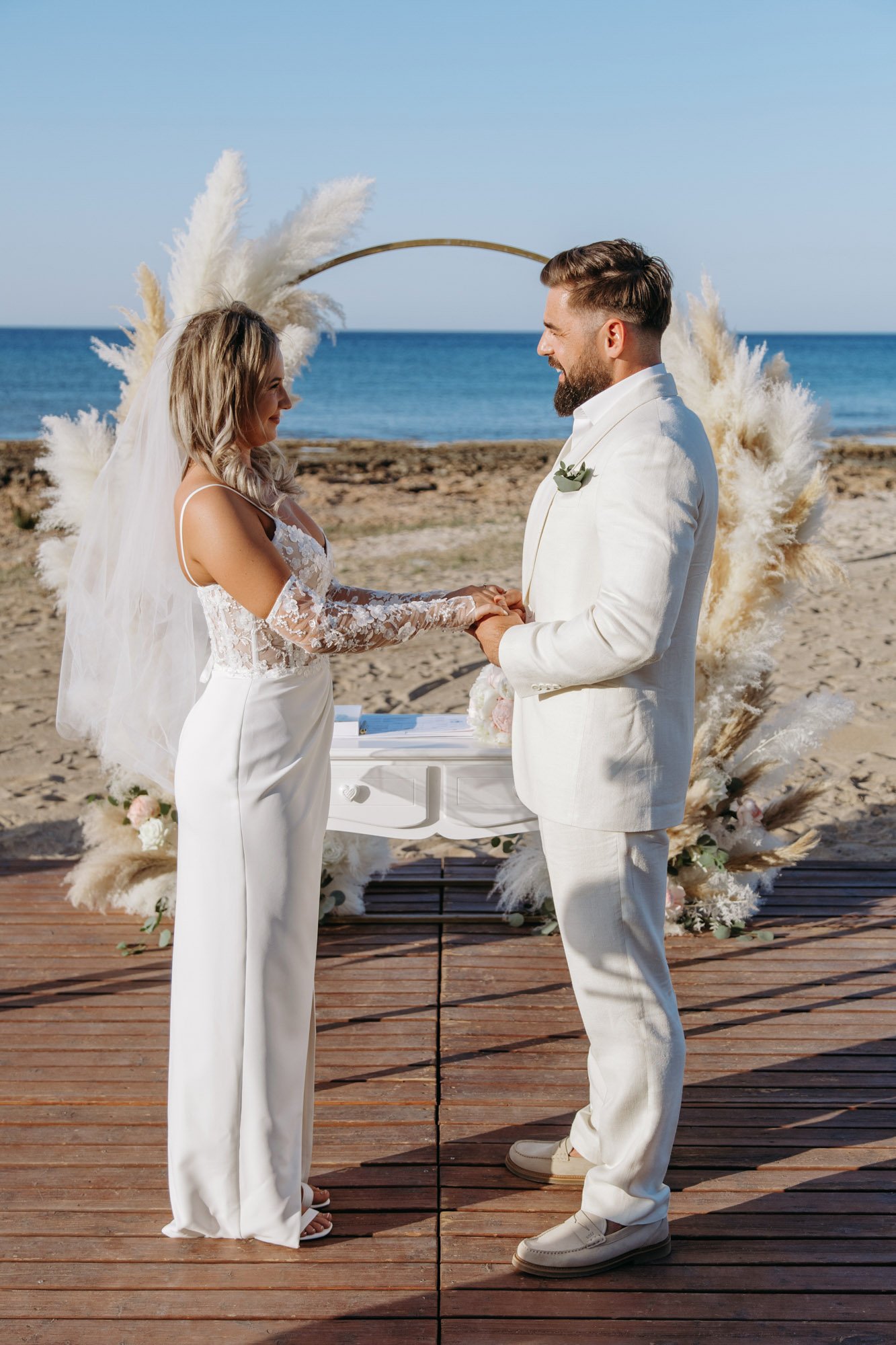 A couple getting married outdoors on a beach, holding hands and smiling at each other, with a floral arch and the ocean in the background.