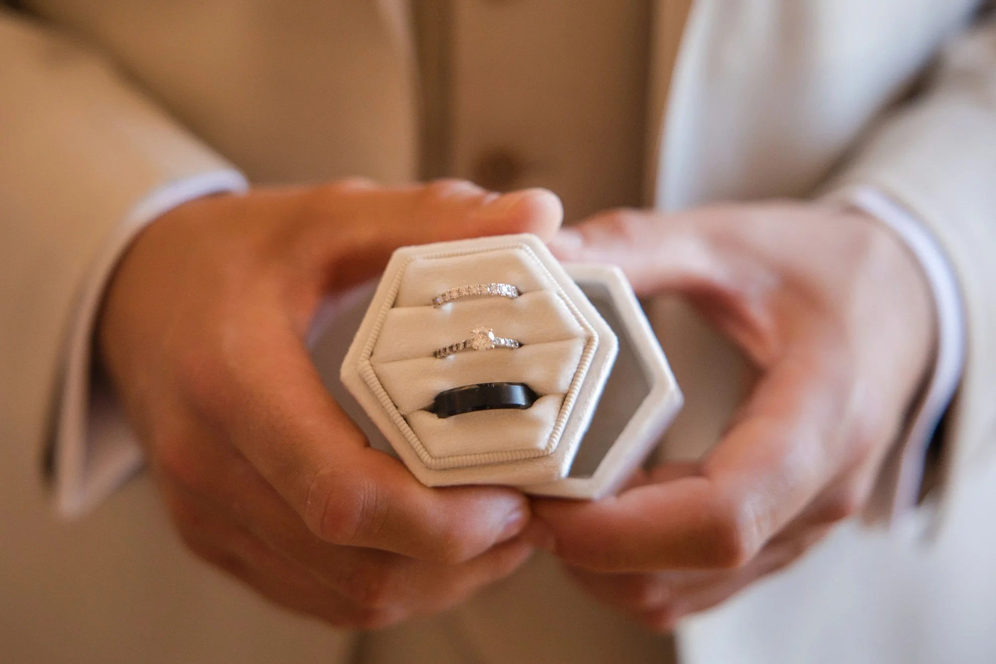 A person in a beige suit holding a white hexagonal jewelry box with three rings inside.