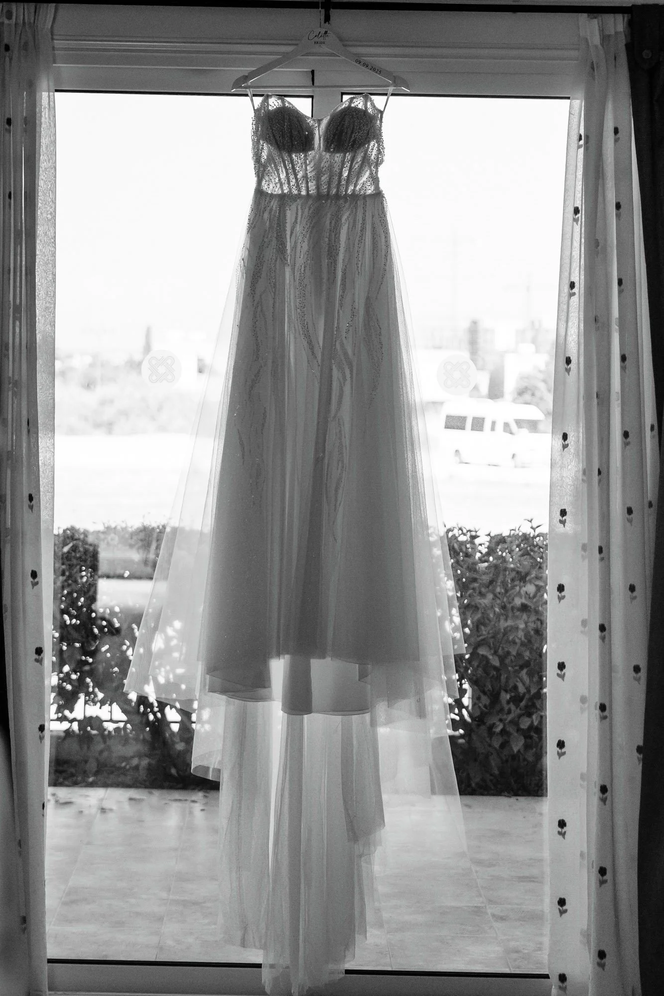 Wedding dress hanging on a hanger in front of a glass door, with curtains and outdoor scenery visible behind.