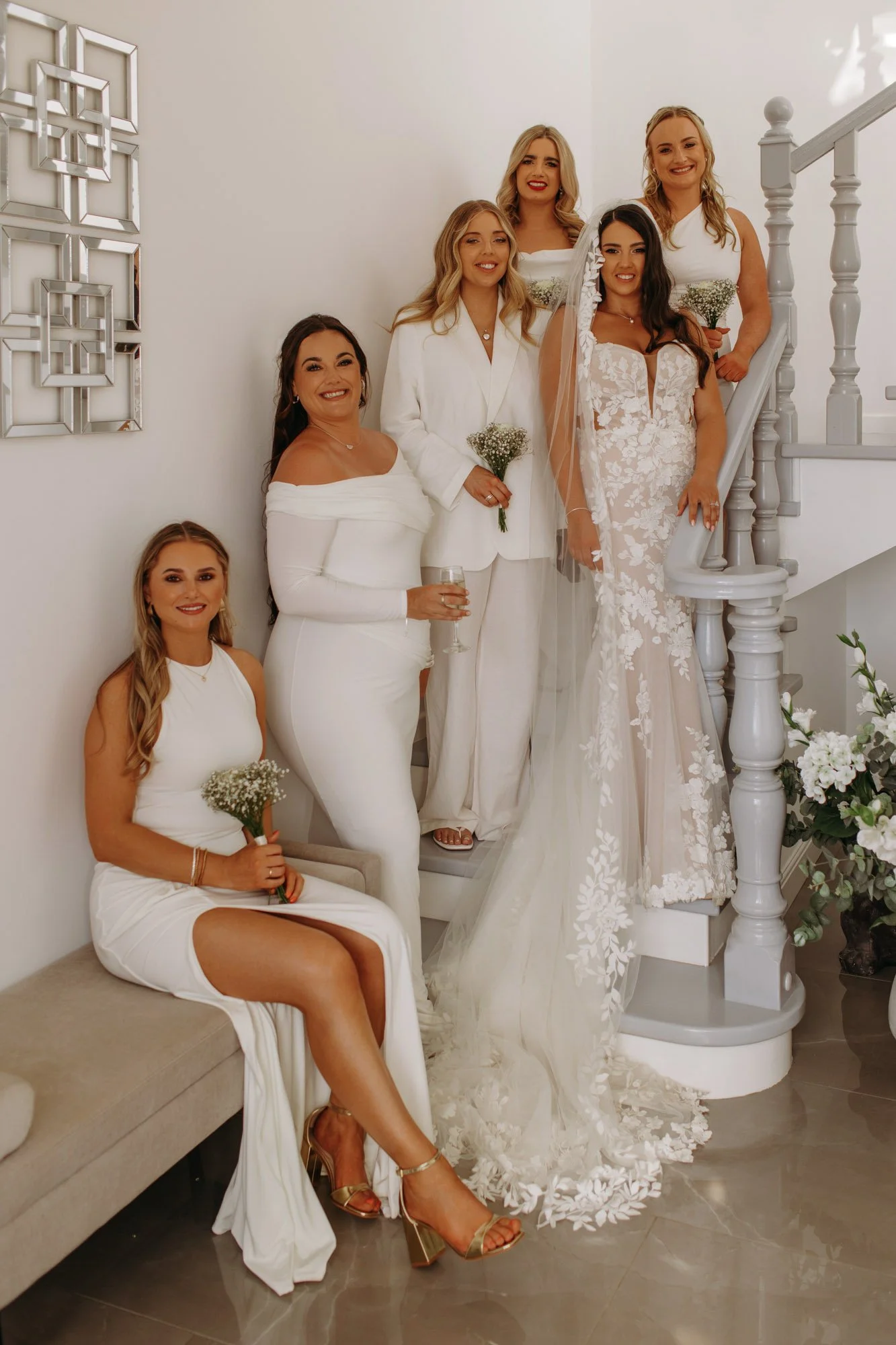 Group of seven women, including a bride in a white wedding dress, posing on a staircase in a home decorated with floral arrangements and modern decor.