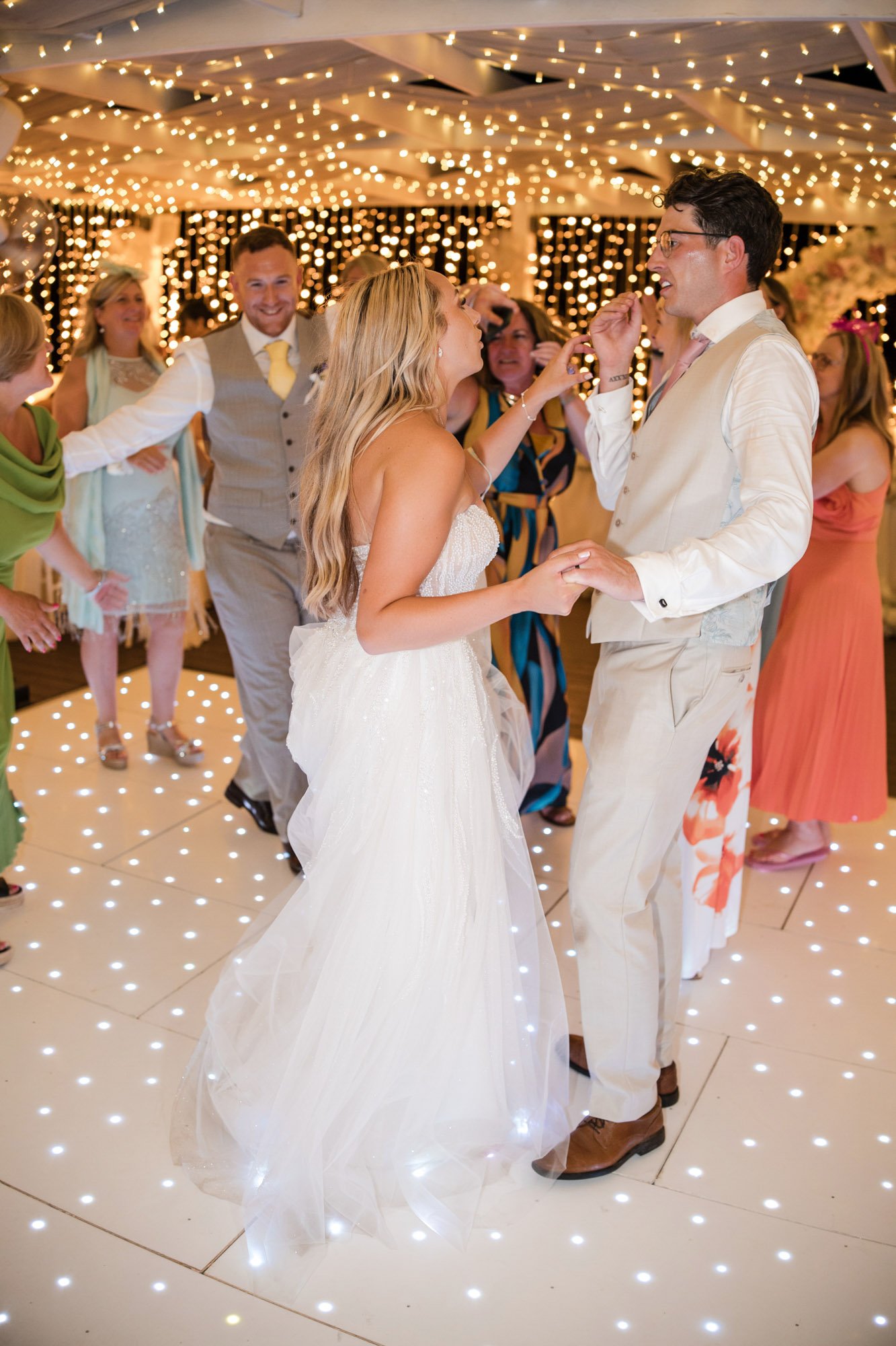 Couple dancing at a wedding reception with string lights and other guests in the background.