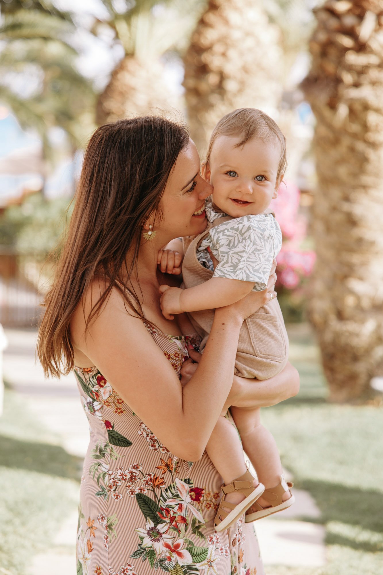 A woman holding a smiling young child outdoors in front of trees with hanging plants.