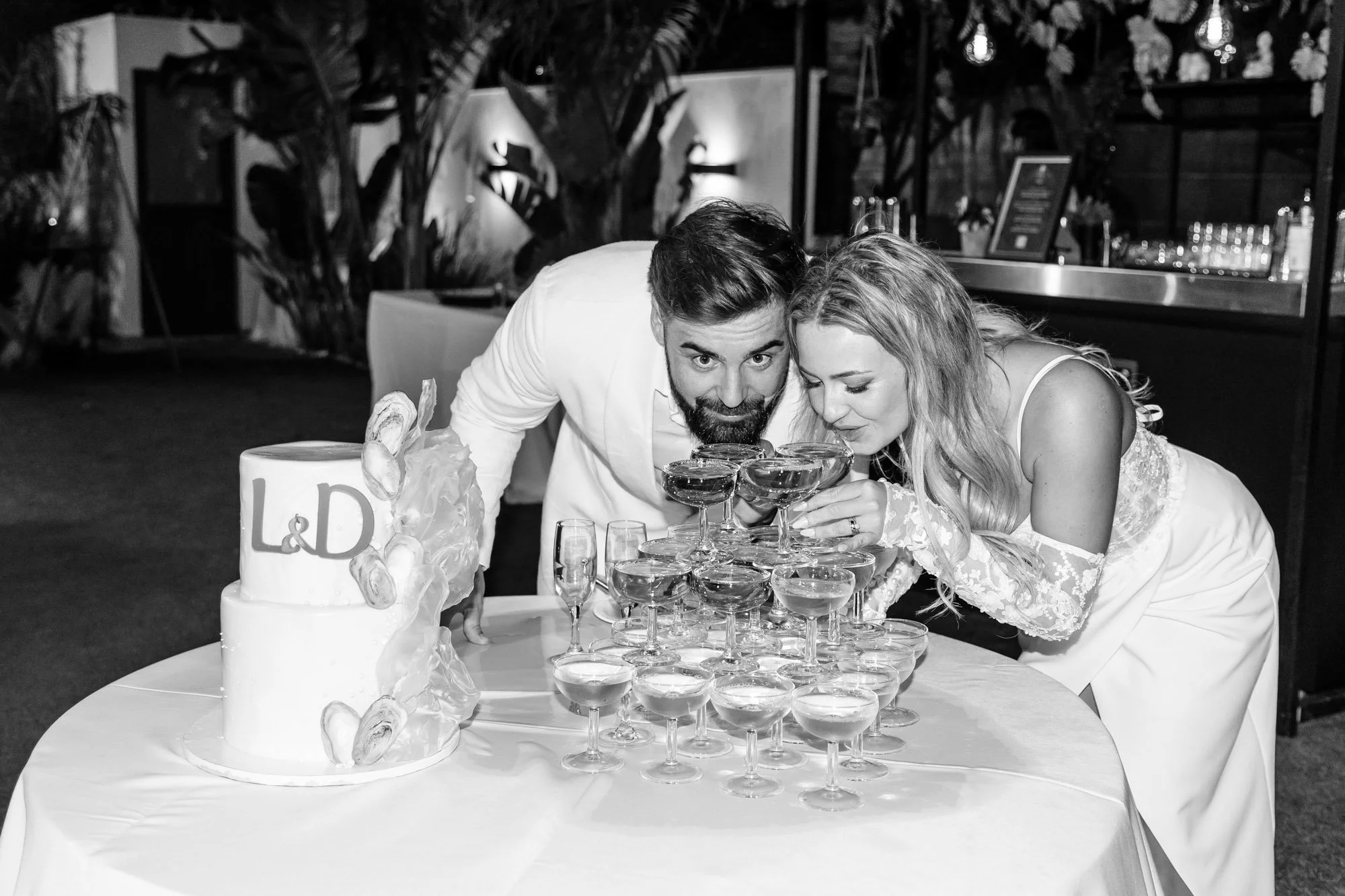 A couple at a wedding, dressed in wedding attire, is being photographed while they are both leaning over a table with a tower of champagne glasses and a wedding cake. They are smiling and looking into the glasses.
