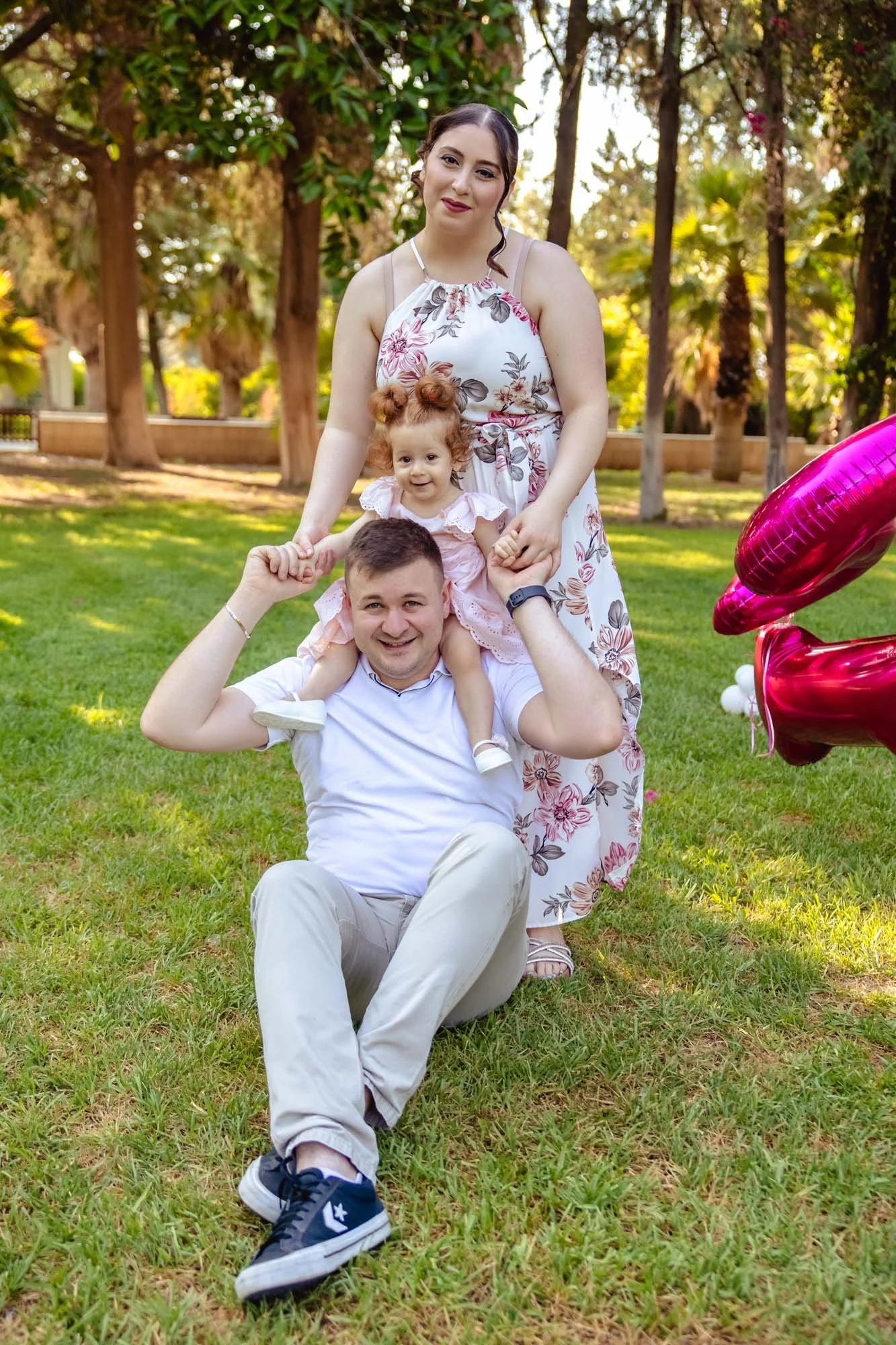 A family of three enjoying an outdoor moment in a park, with trees and grass in the background, and pink heart-shaped balloons on the right.
