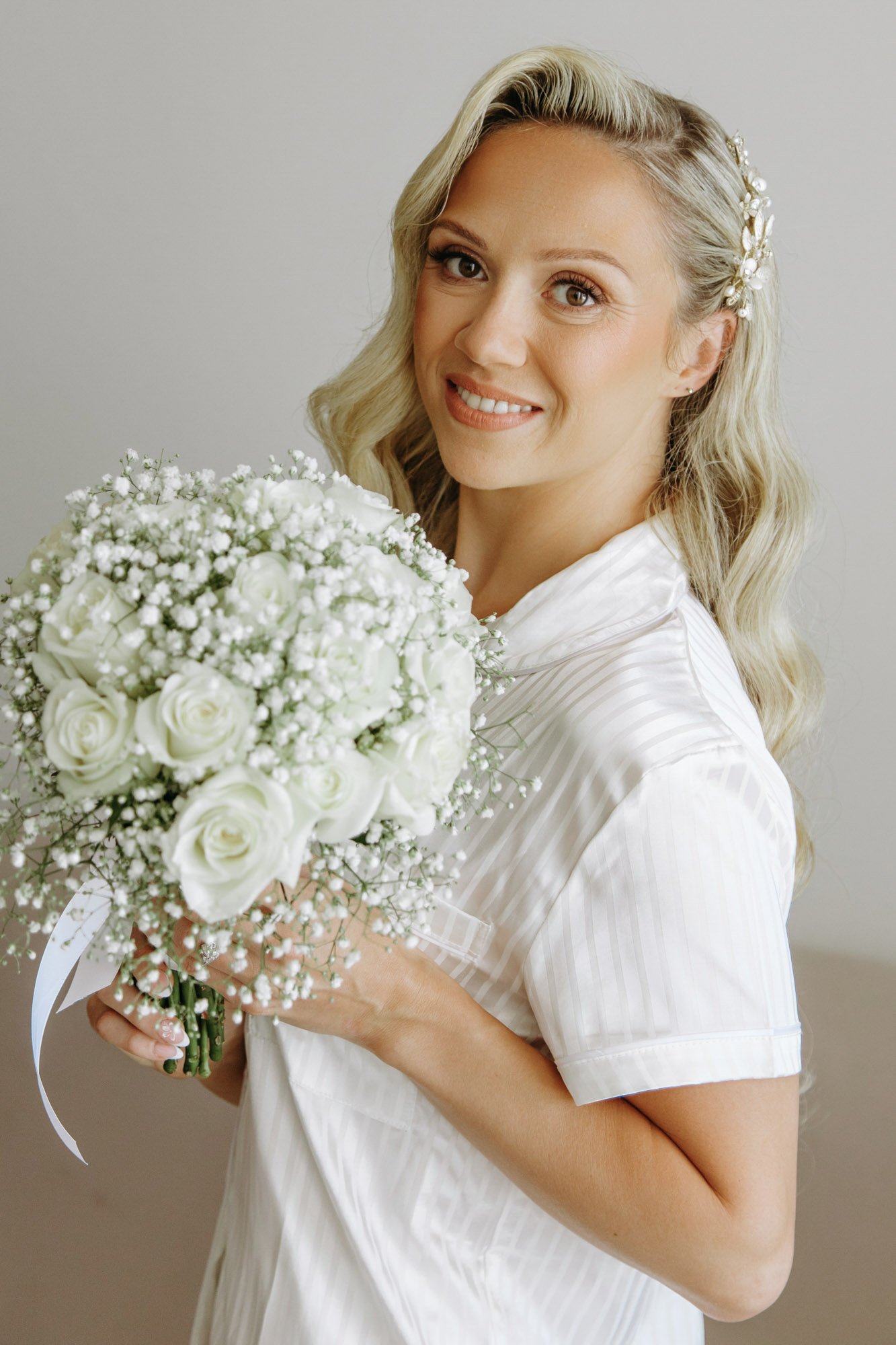 A woman with long wavy blonde hair, wearing a white striped shirt, is holding a bouquet of white roses and baby's breath. She is smiling and has a pearl hair accessory.