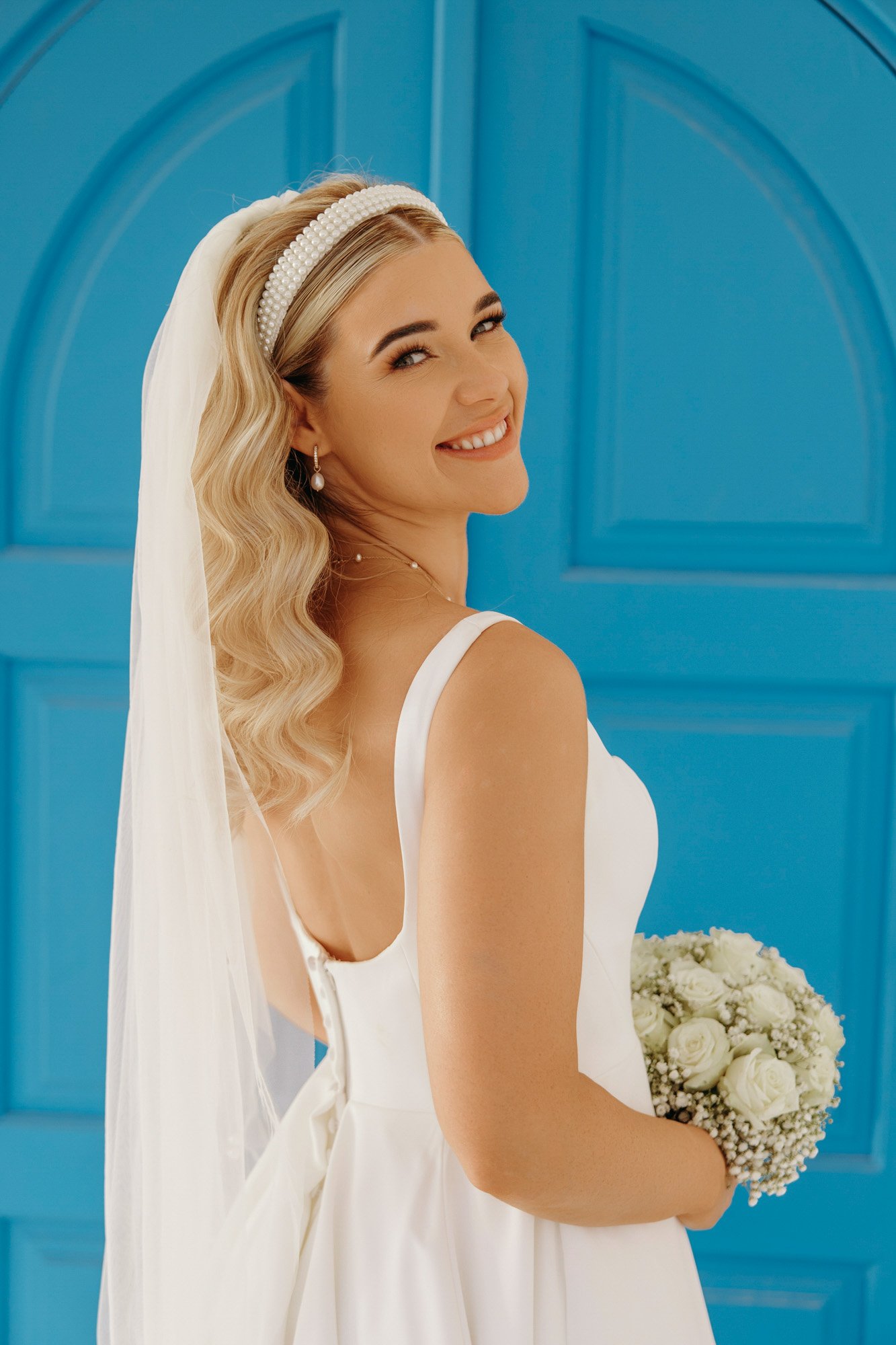 A smiling bride dressed in a white gown, holding a bouquet of white roses, standing in front of a bright blue background.