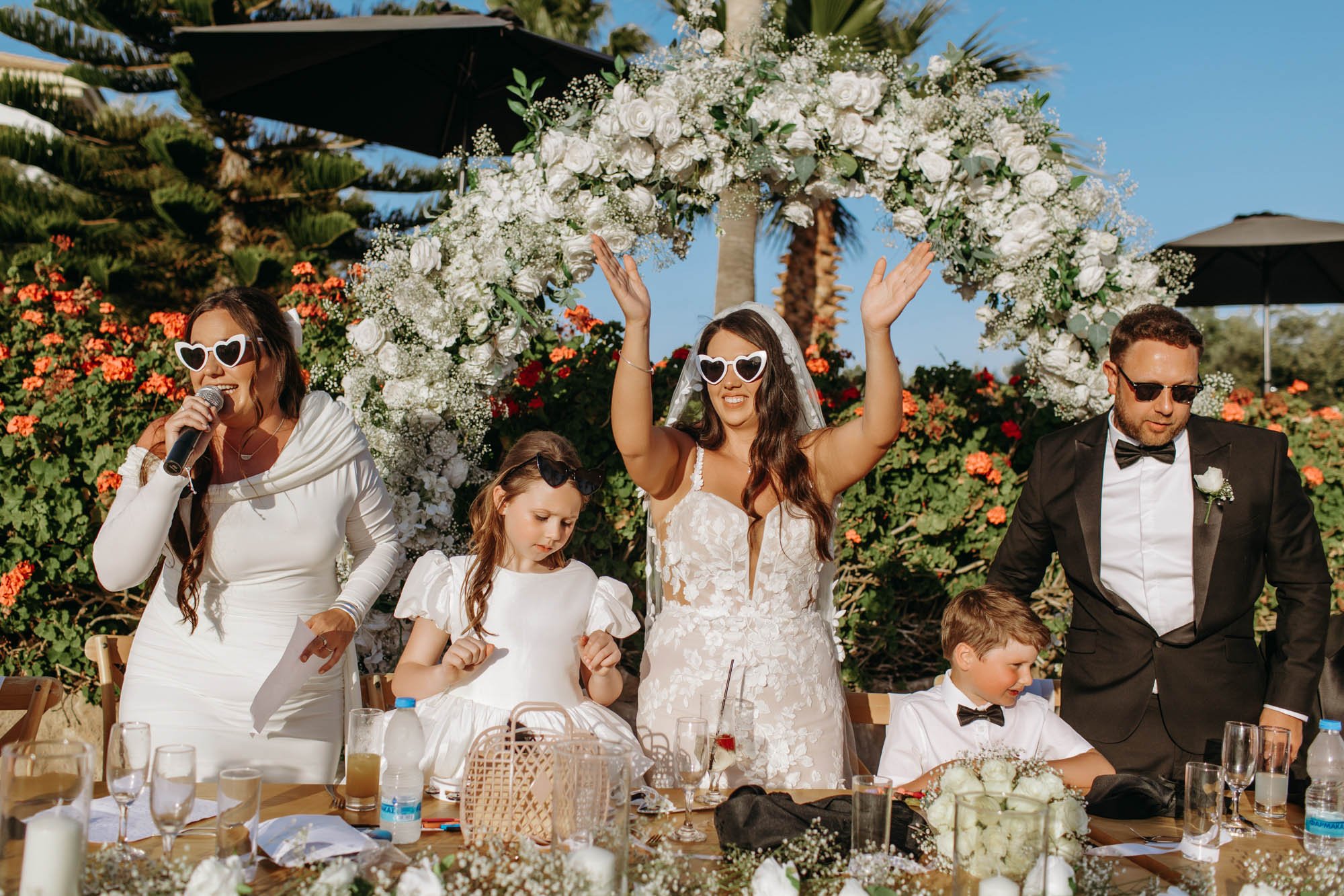 Couples and children celebrating at a wedding reception outdoors under a floral arch with white roses and greenery, with palm trees and a blue sky in the background.