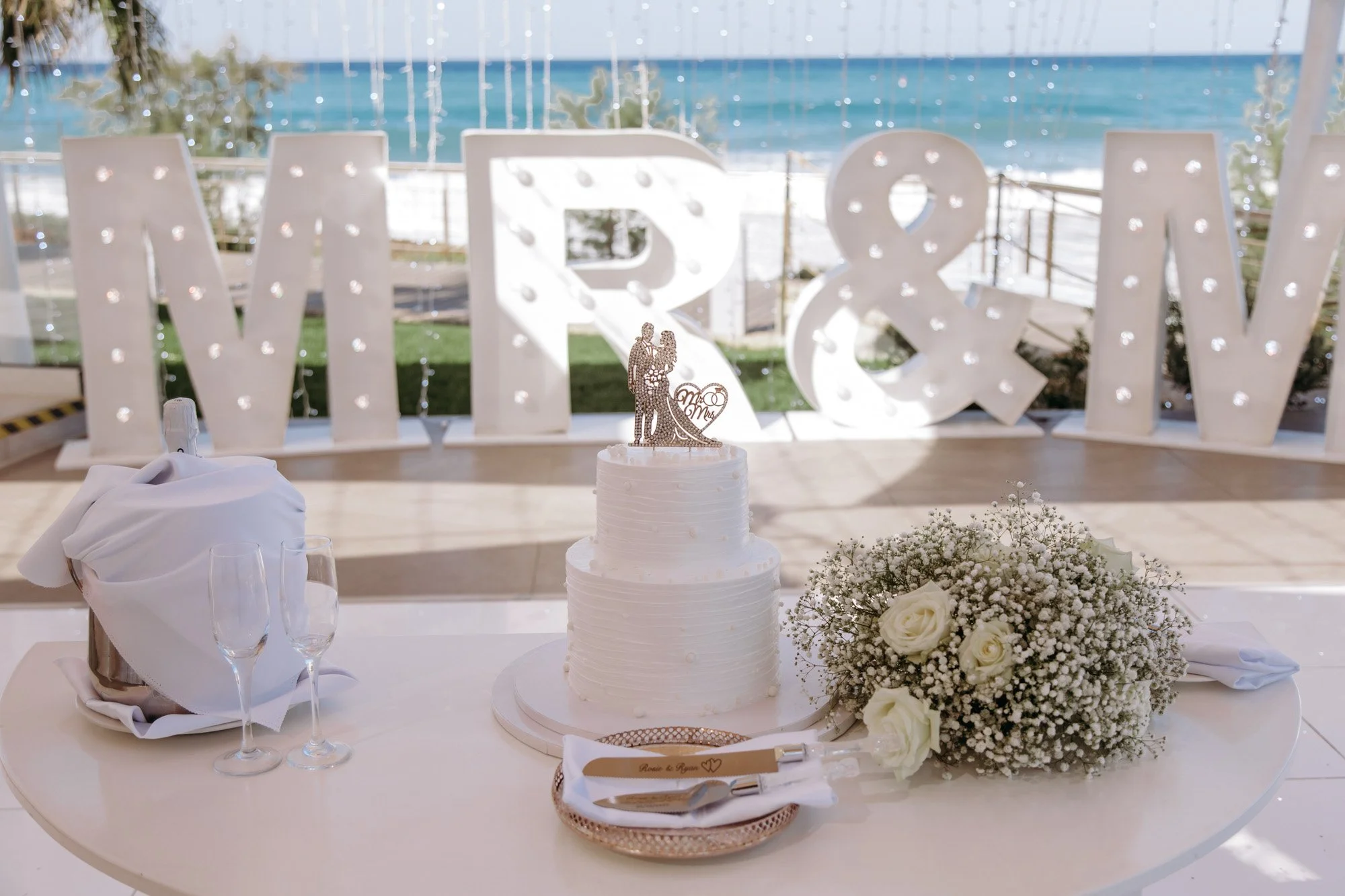 Wedding cake decorated with a bride and groom topper, second tier with white flowers, and a bouquet of white roses and baby's breath on the table, with large illuminated letters 'M', 'R', '&', 'W' in the background overlooking the ocean.