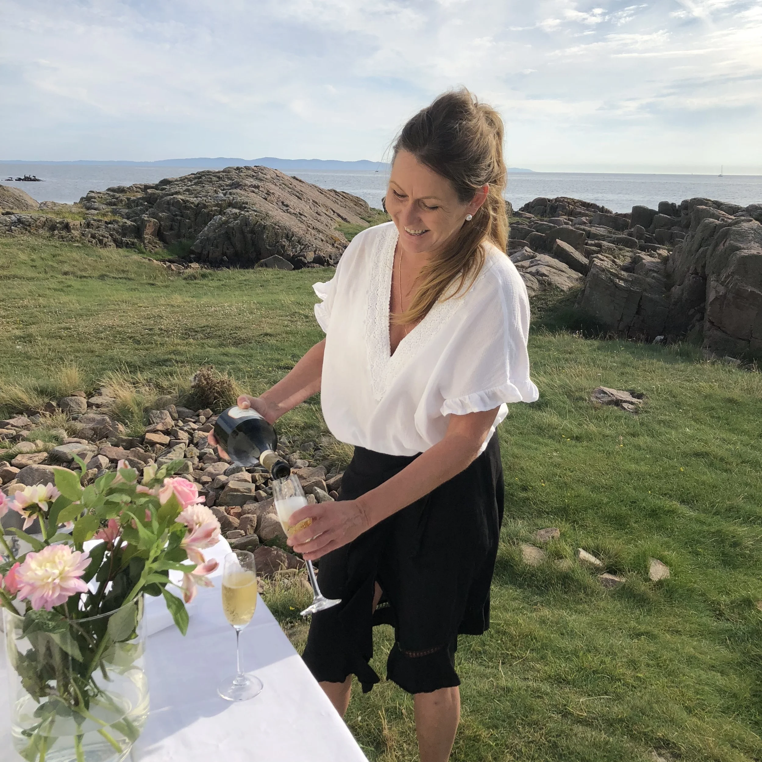 En kvinna i vit blus och svarta byxor som fyller ett glas med champagne vid ett utebord med blommor, hav och klippor i bakgrunden.