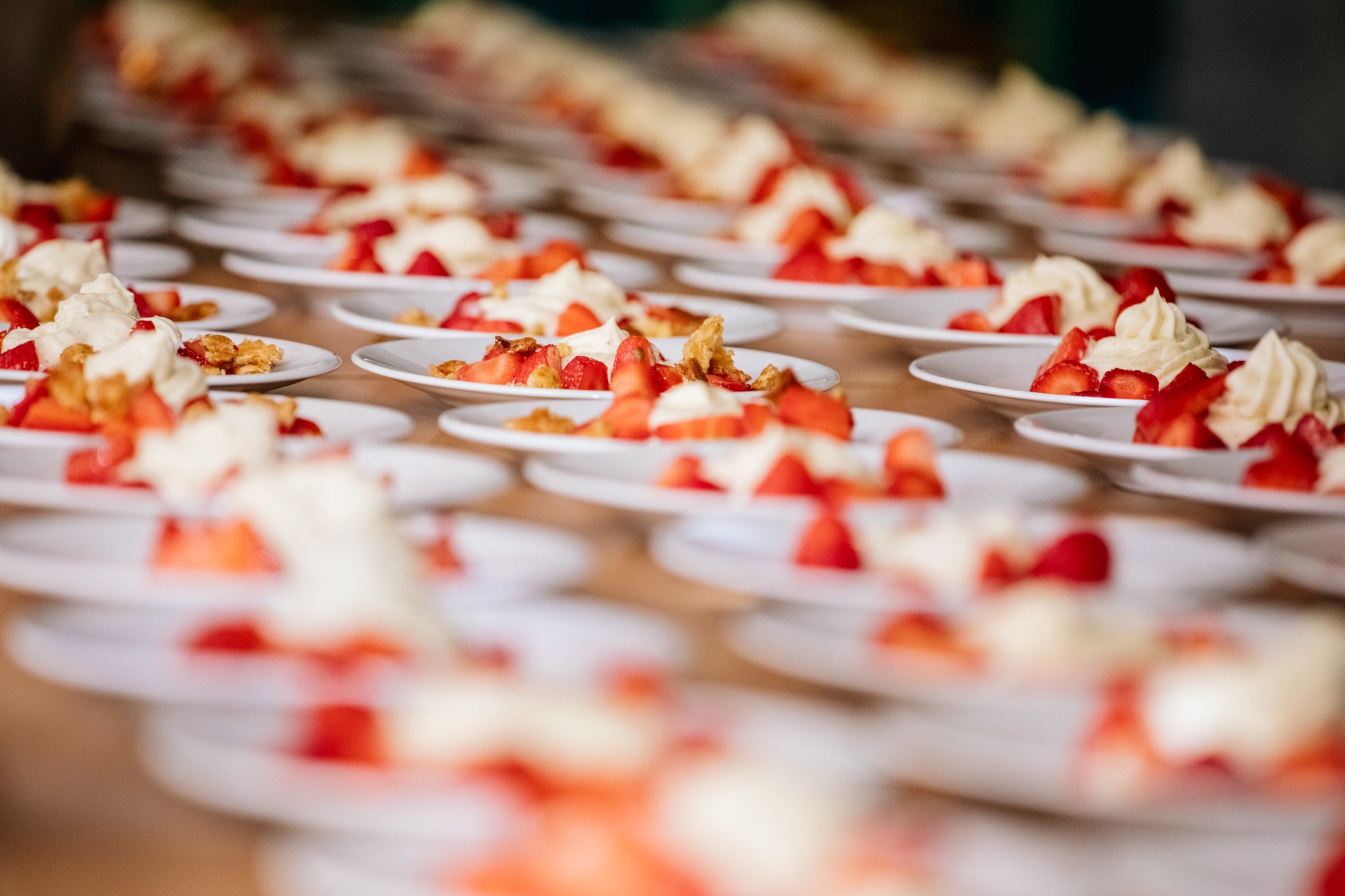 Flera små uppläggningar av jordgubbar, grädde och krossad kex på vita tallrikar, arrangerade i rader.